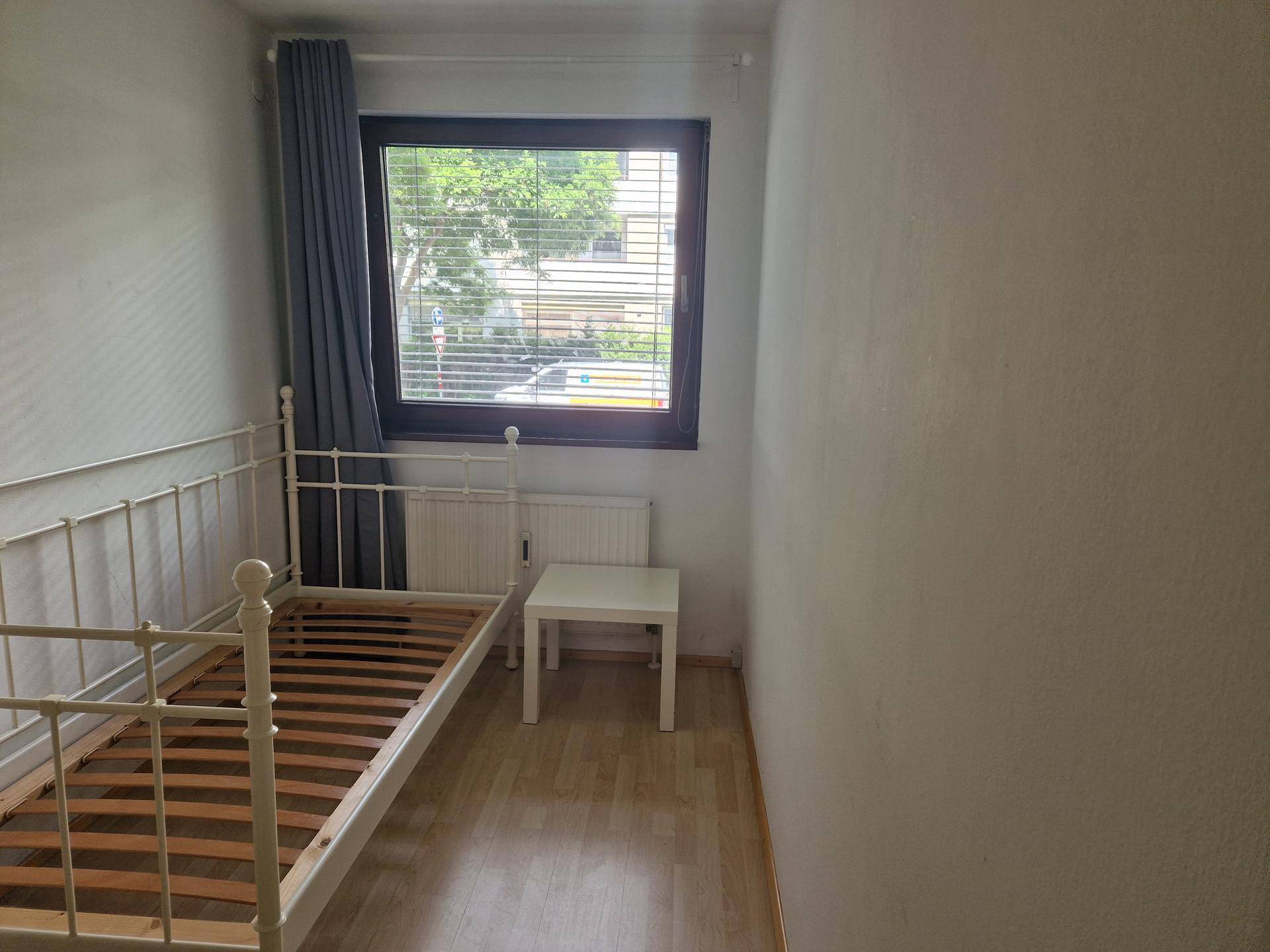 Helles Schlafzimmer mit weißem Metallbett, kleinem Beistelltisch und Fenster mit Jalousien.