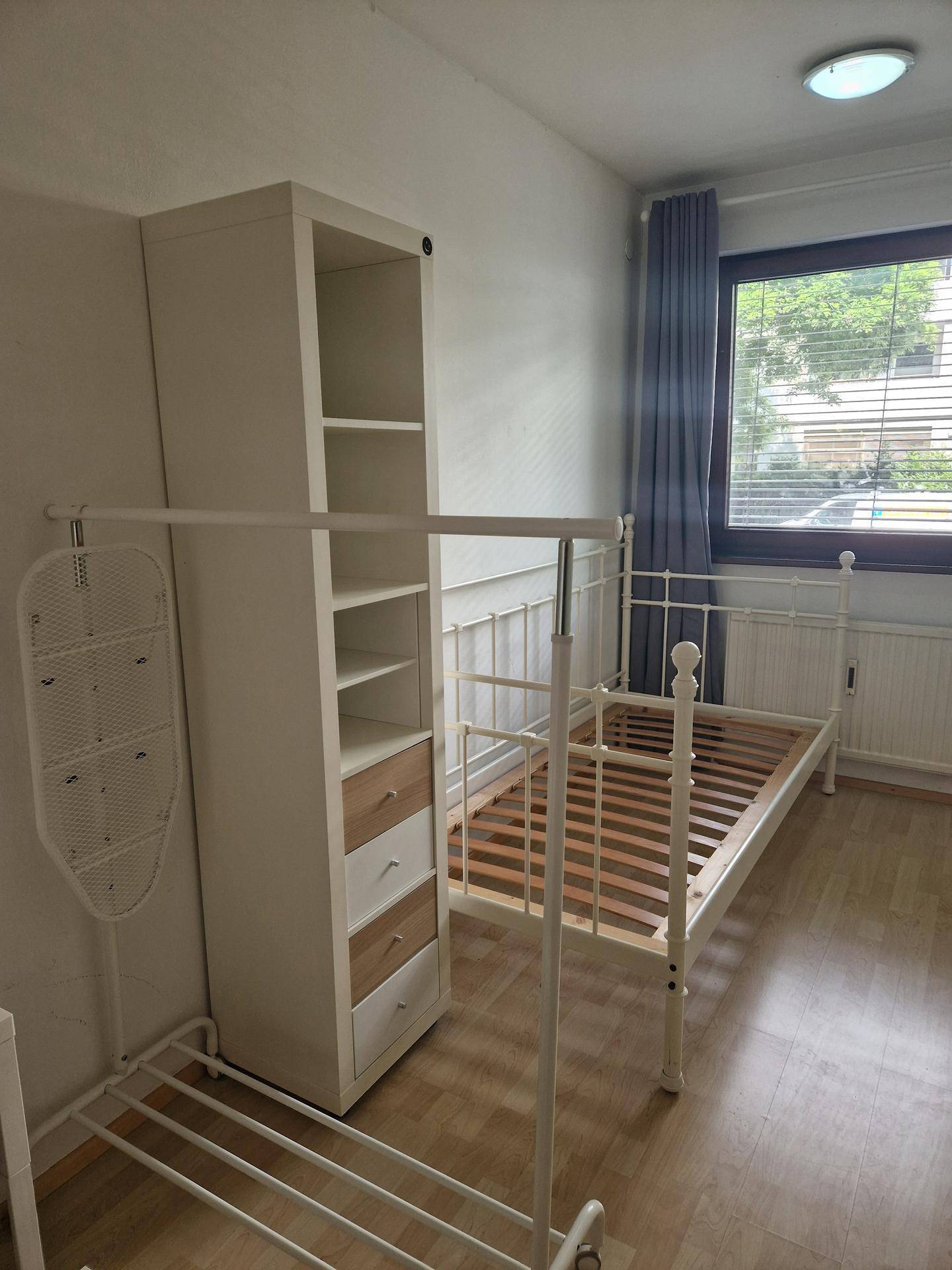 Schlafzimmer mit weißem Metallbett, hohem Regal und Fenster mit blauen Vorhängen.