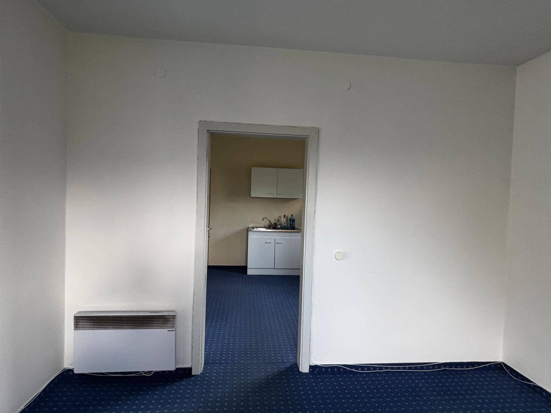 Leerer Büroraum mit blauem Teppichboden und Durchgang zu einer kleinen Küchenzeile.