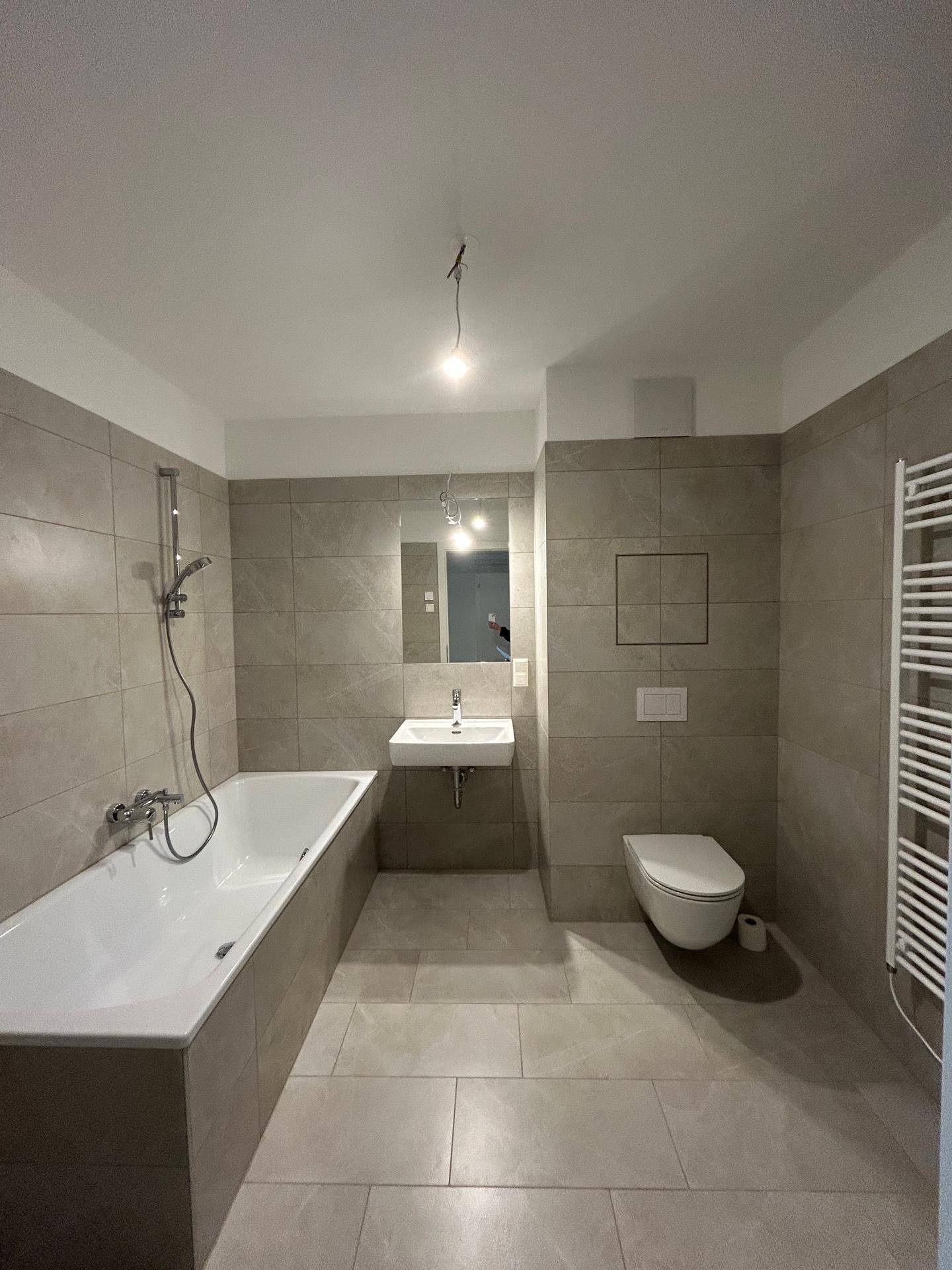 Modernes Badezimmer mit Badewanne, Wand-WC und großformatigen Fliesen, ausgestattet mit Handtuchheizkörper.