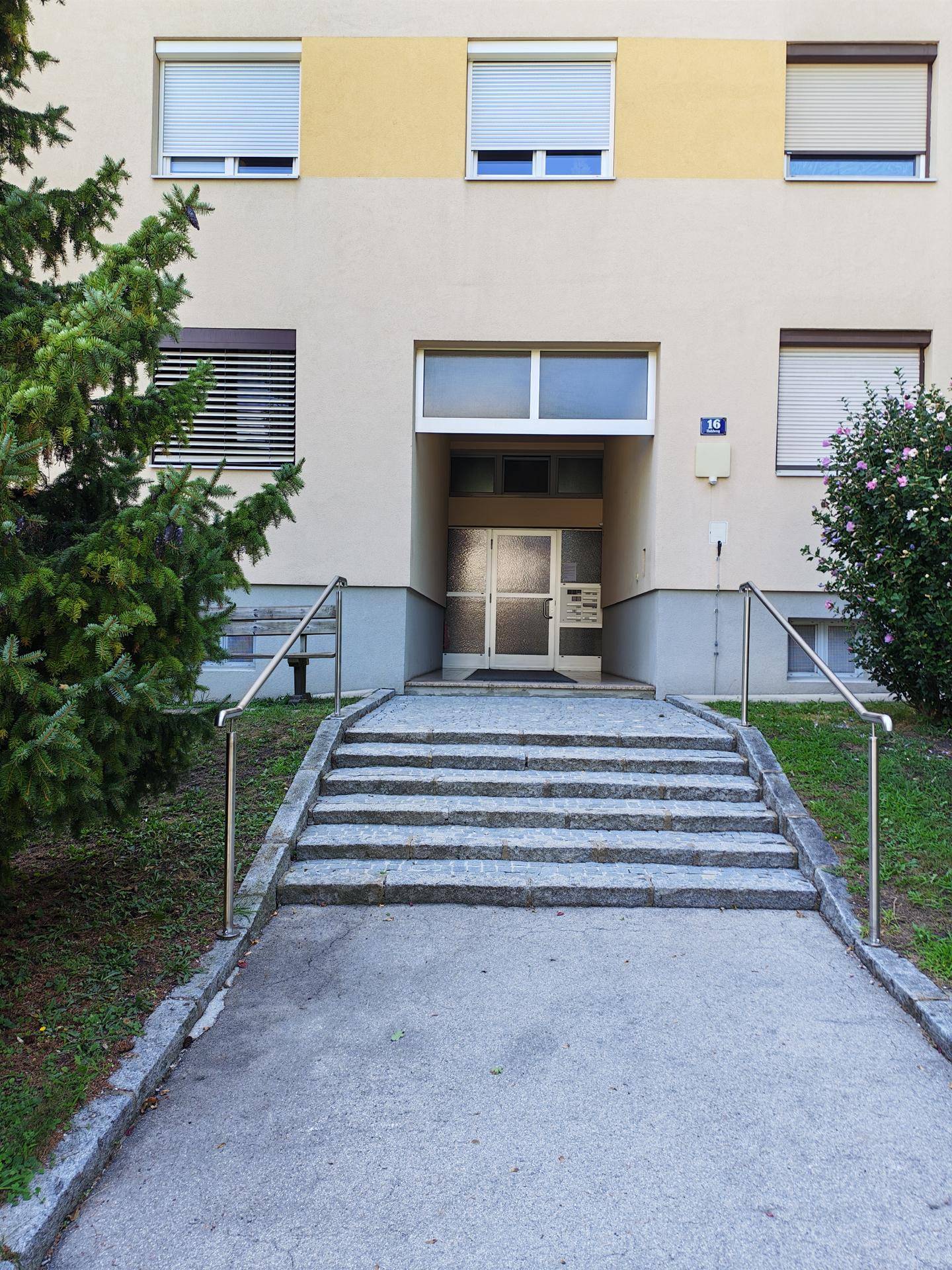 Eingangsbereich eines Mehrfamilienhauses mit Steintreppe, Geländer und Rollläden an den Fenstern.