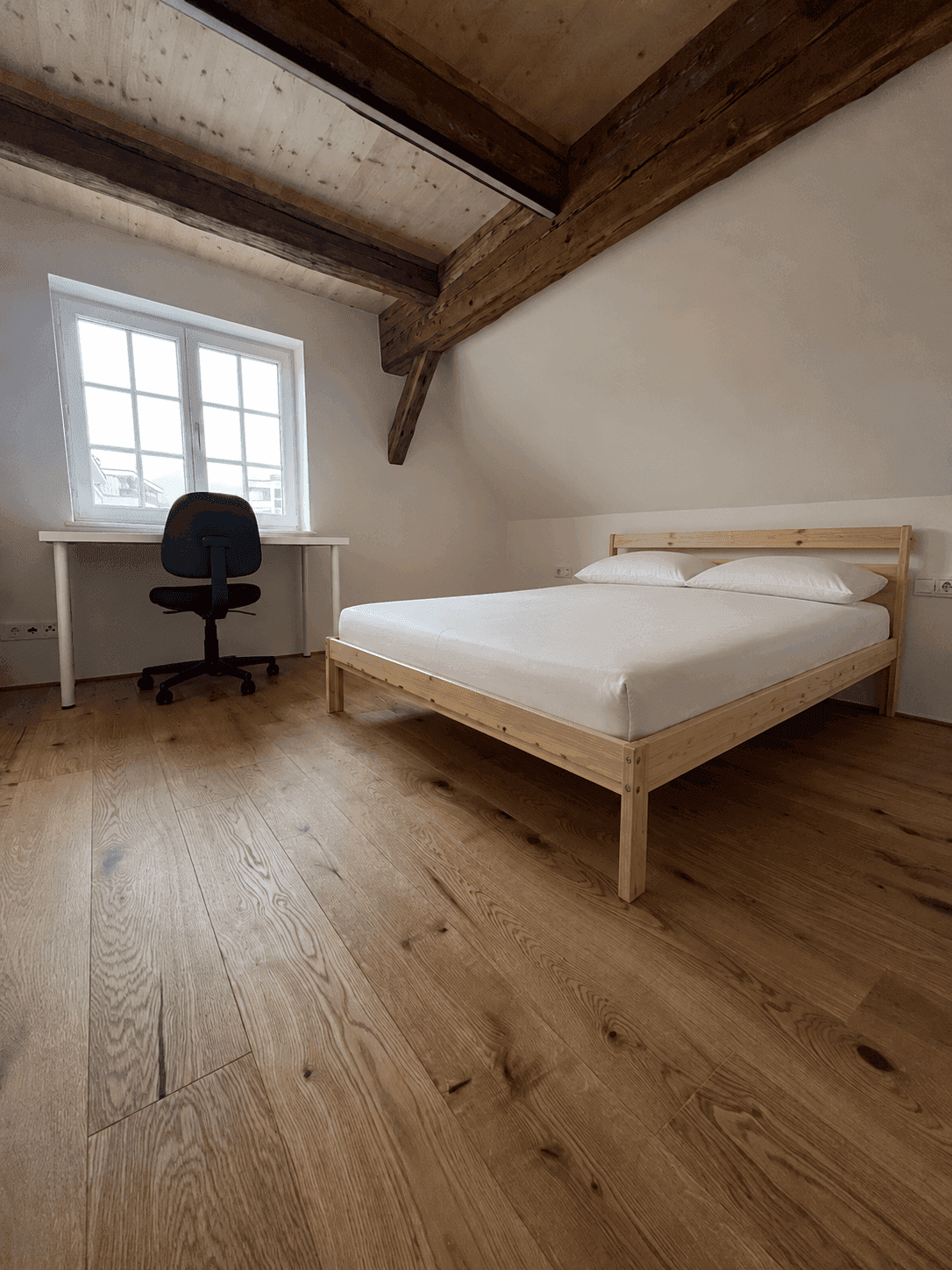 Helles Schlafzimmer mit Holzbett, Schreibtisch und Fenster im Dachgeschoss.