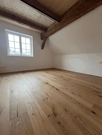 Helles Schlafzimmer mit Holzboden, sichtbaren Holzbalken und einem großen Fenster.