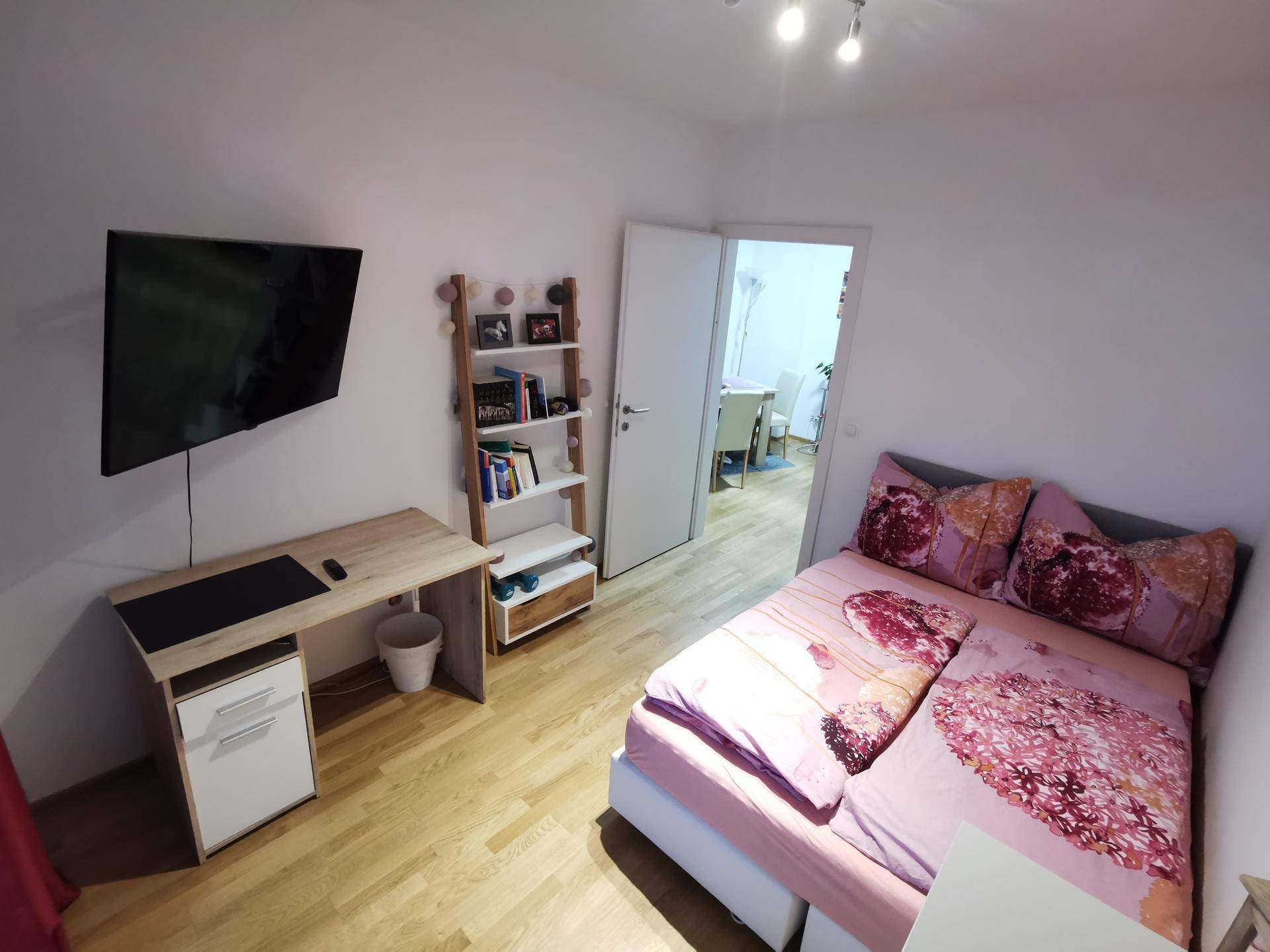 Gemütliches Schlafzimmer mit Holzboden, einem Bett, Schreibtisch und einem Regal, ideal für Ruhe und Arbeit.