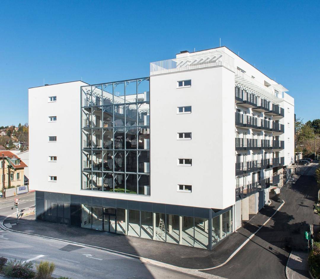 Modernes Wohngebäude mit weißer Fassade, großen Glasflächen und mehreren Balkonen unter blauem Himmel.