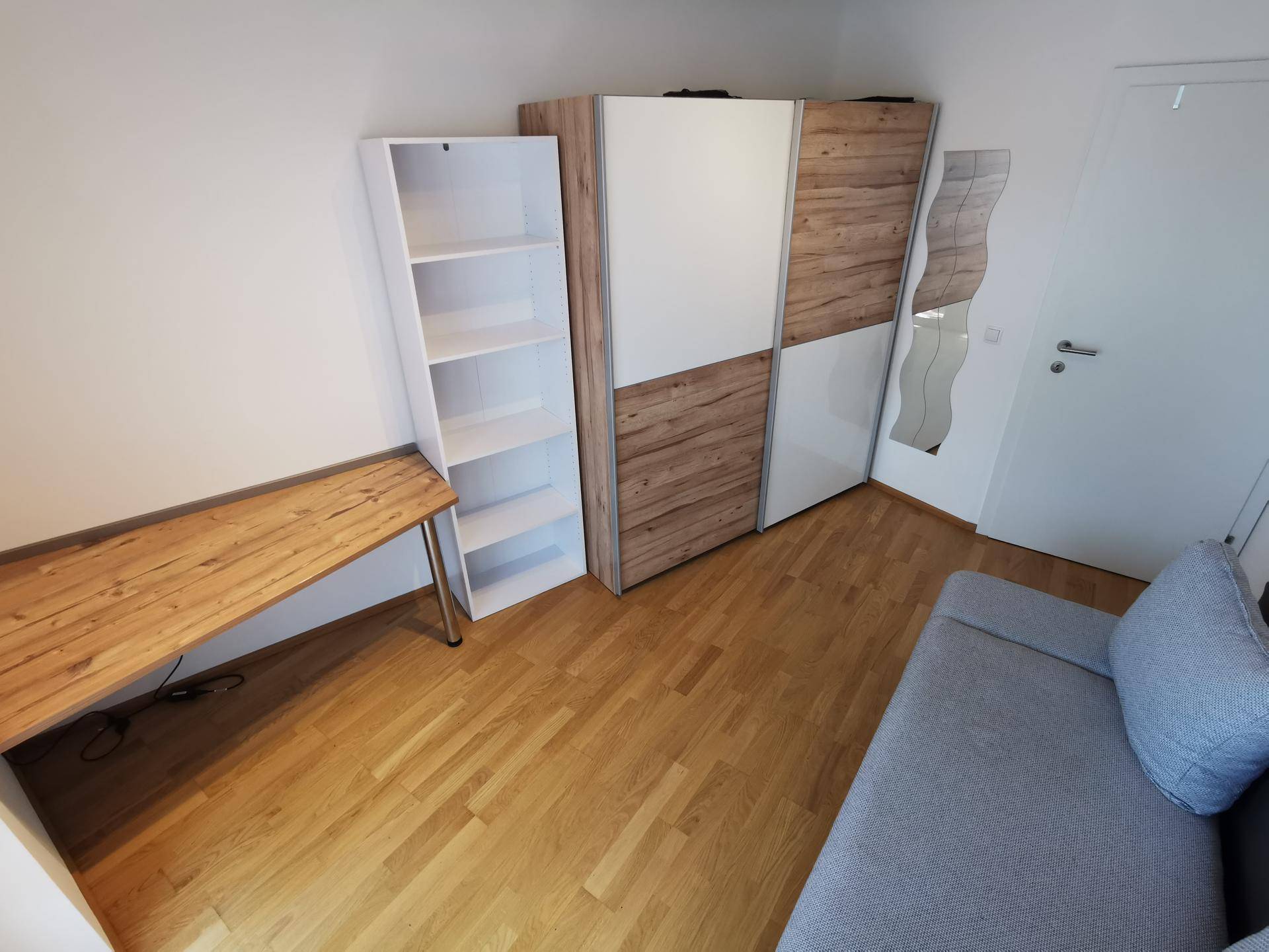 Helles Zimmer mit Holzboden, einem bequemen Sofa, Schreibtisch und einem modernen Kleiderschrank.