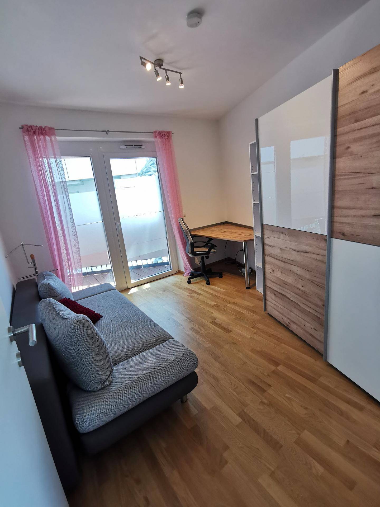 Freundliches Zimmer mit Sofa, Schreibtisch und Zugang zu einem Balkon, geschmückt mit rosa Vorhängen.