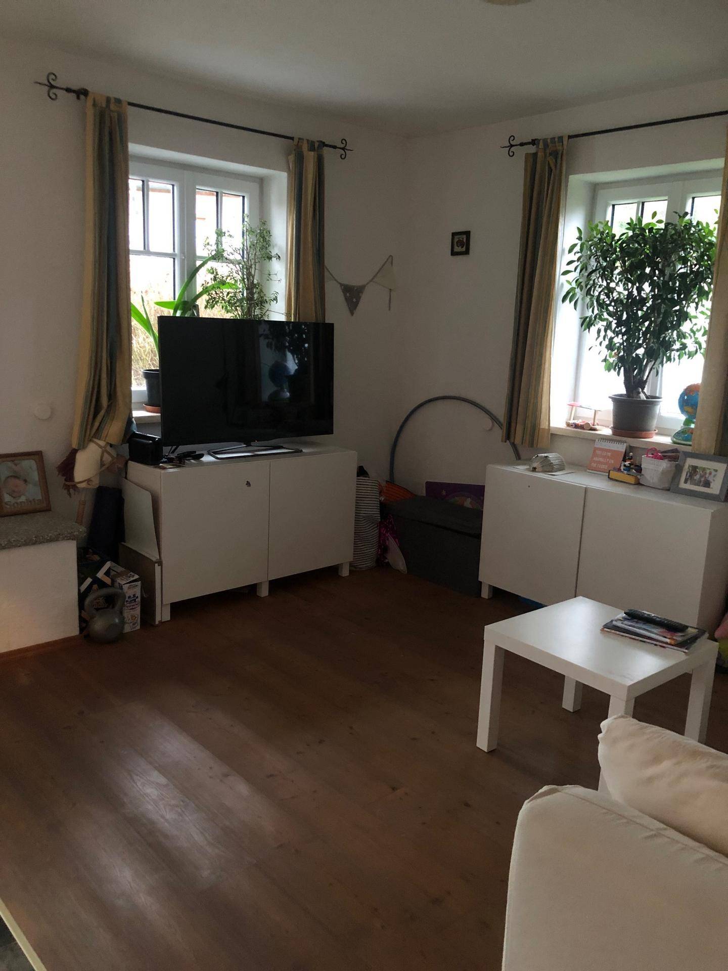 Helles Wohnzimmer mit Holzboden, zwei Fenstern und schlichten weißen Möbeln.
