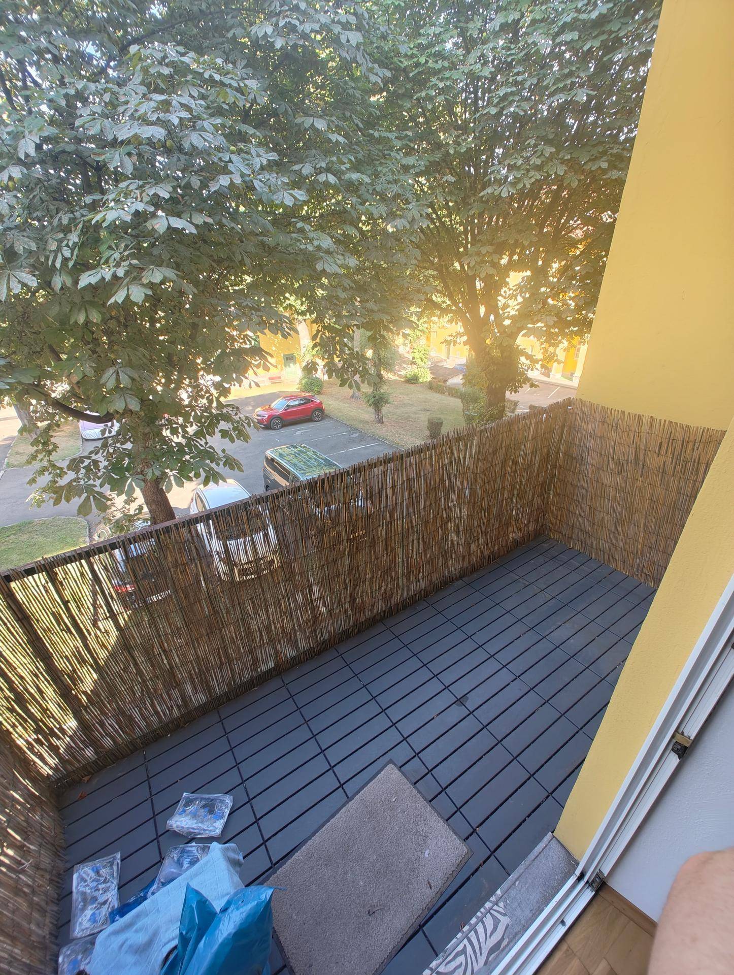 Balkon mit Holzbodenfliesen, Sichtschutz und Blick auf Bäume und Parkplätze.