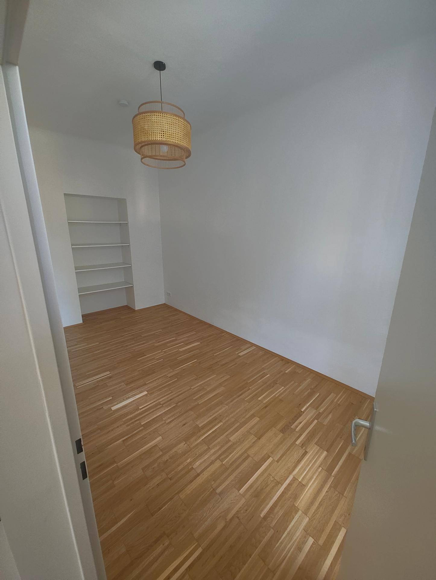 Leeres Zimmer mit Parkettboden, Einbauregal und einer modernen Deckenleuchte.