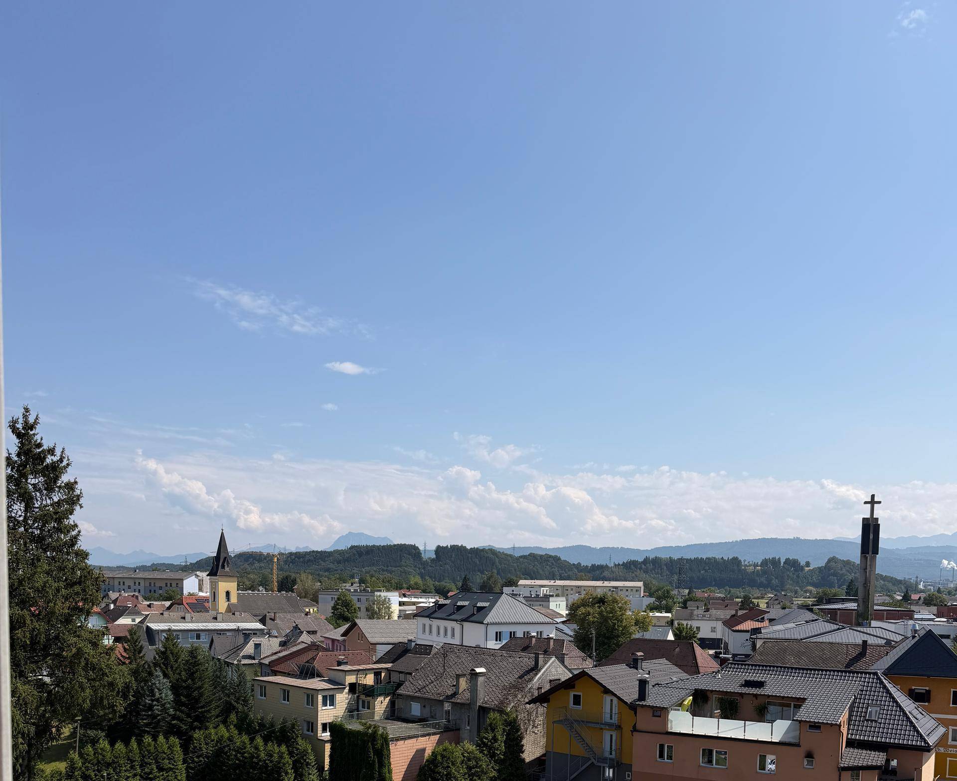 Panoramablick über eine Stadt mit Kirchturm und Bergen am Horizont unter blauem Himmel.