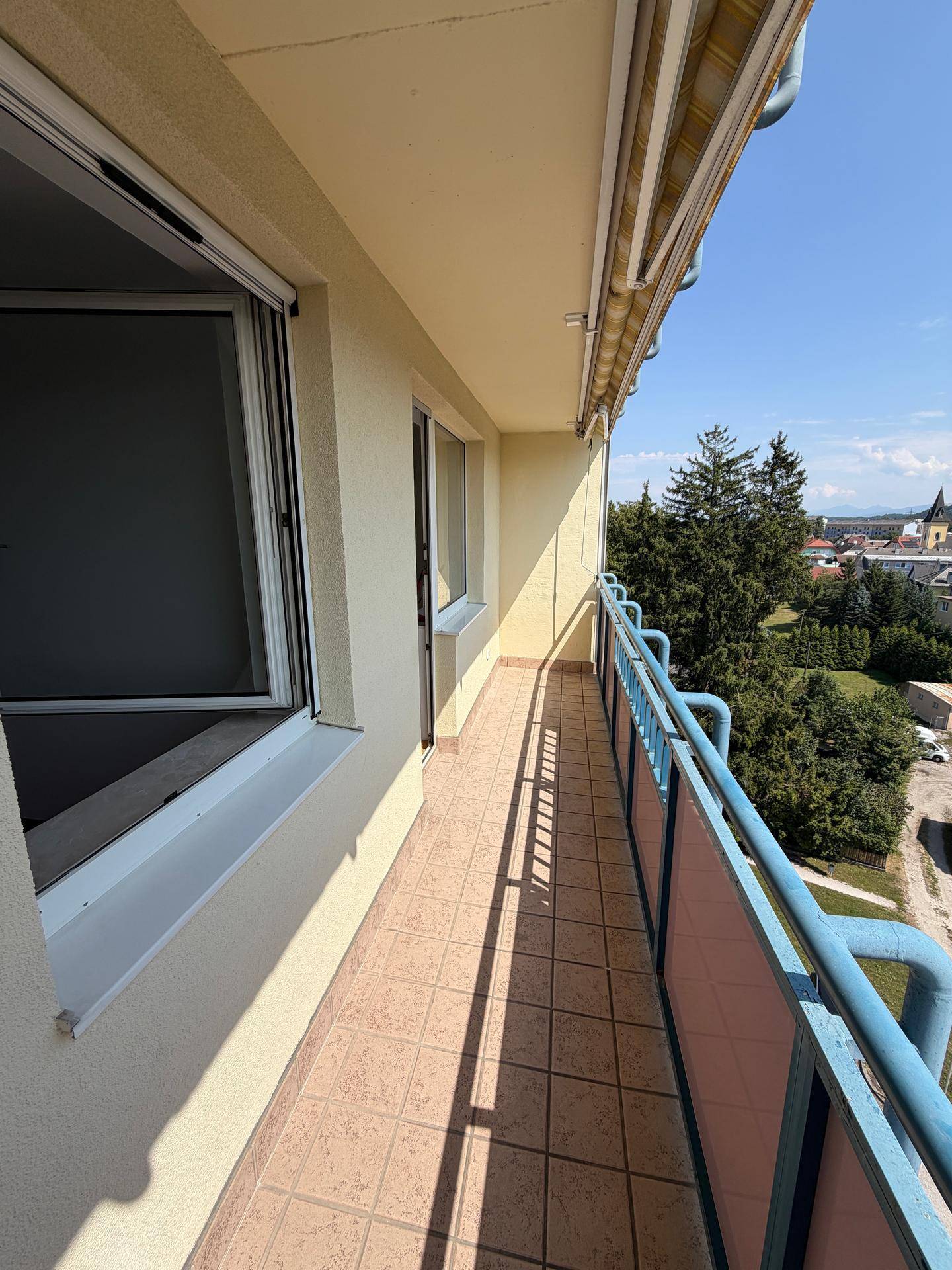 Langer Balkon mit Fliesenboden und blauem Geländer, der einen Blick auf die Umgebung bietet.