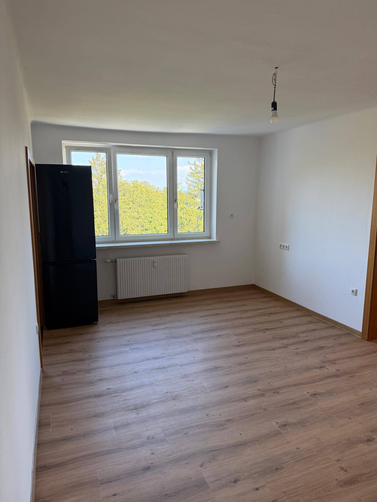 Leerer Raum mit Laminatboden, großem Fenster und einem freistehenden Kühlschrank.