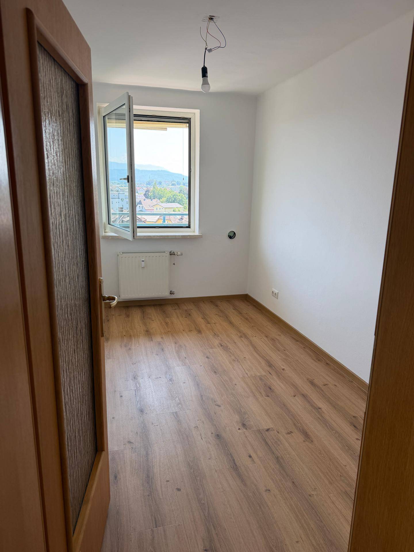 Leeres Zimmer mit Laminatboden und einem Fenster, das viel Tageslicht hereinlässt.