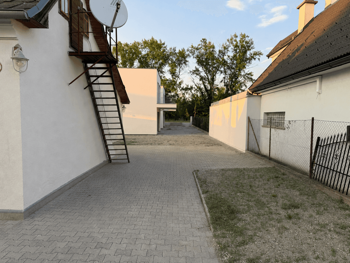Gepflasterte Einfahrt zwischen Gebäuden mit Satellitenschüssel an der Fassade, bei Abendlicht.