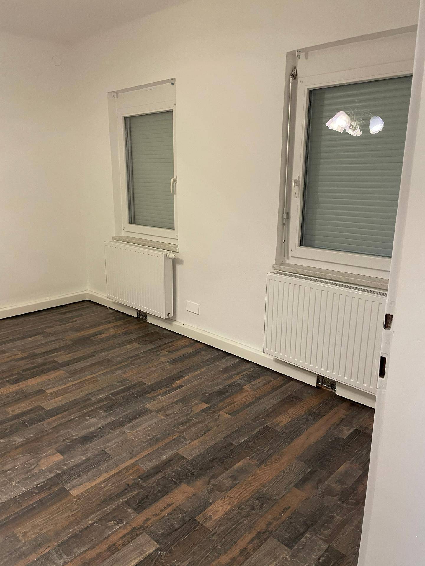 Leeres Schlafzimmer mit dunklem Laminatboden, weißen Wänden und zwei Fenstern mit Heizkörpern darunter.