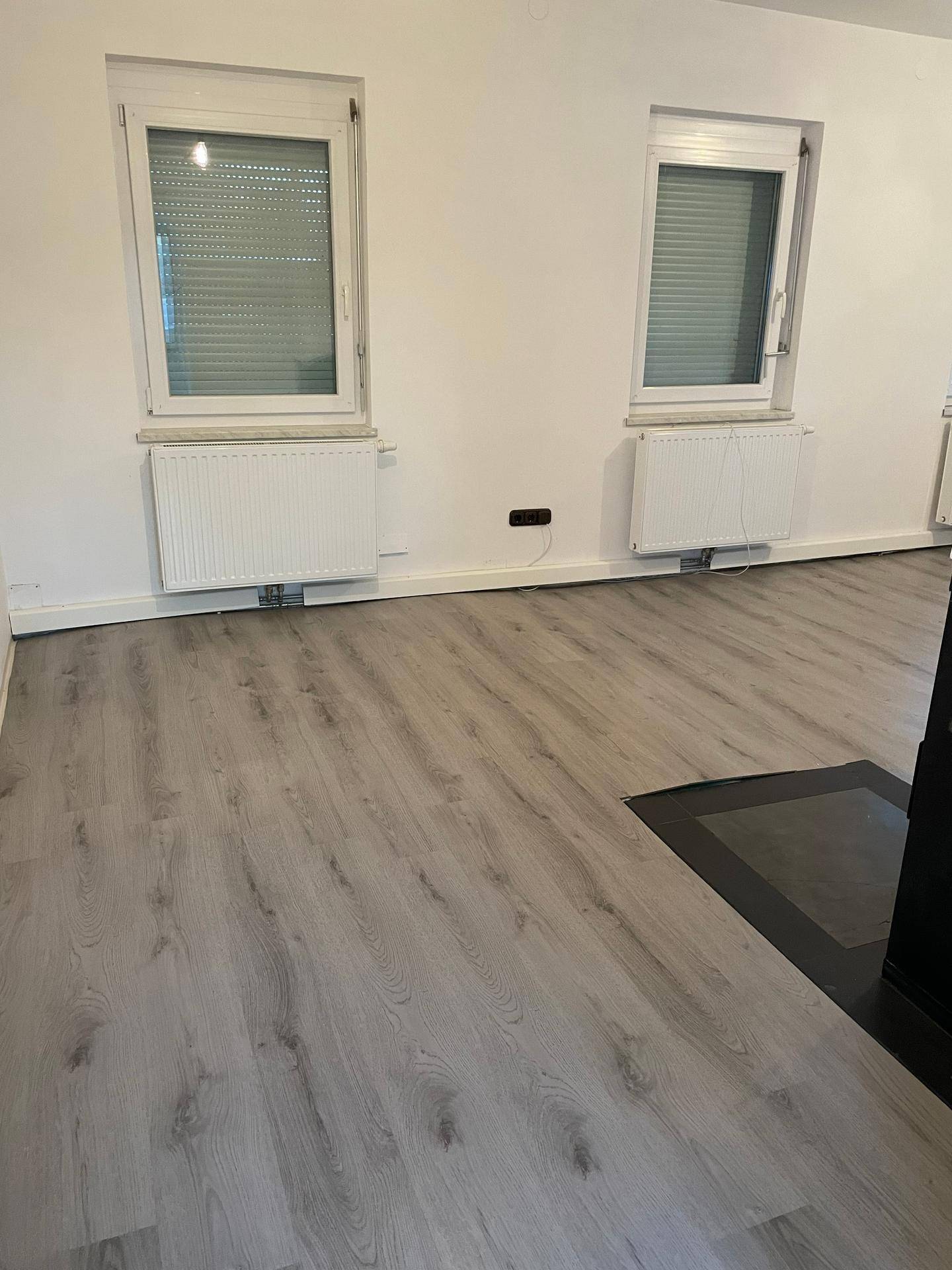 Helles Wohnzimmer mit hellem Laminatboden, weißen Wänden und zwei Fenstern, vorbereitet für einen Ofen.