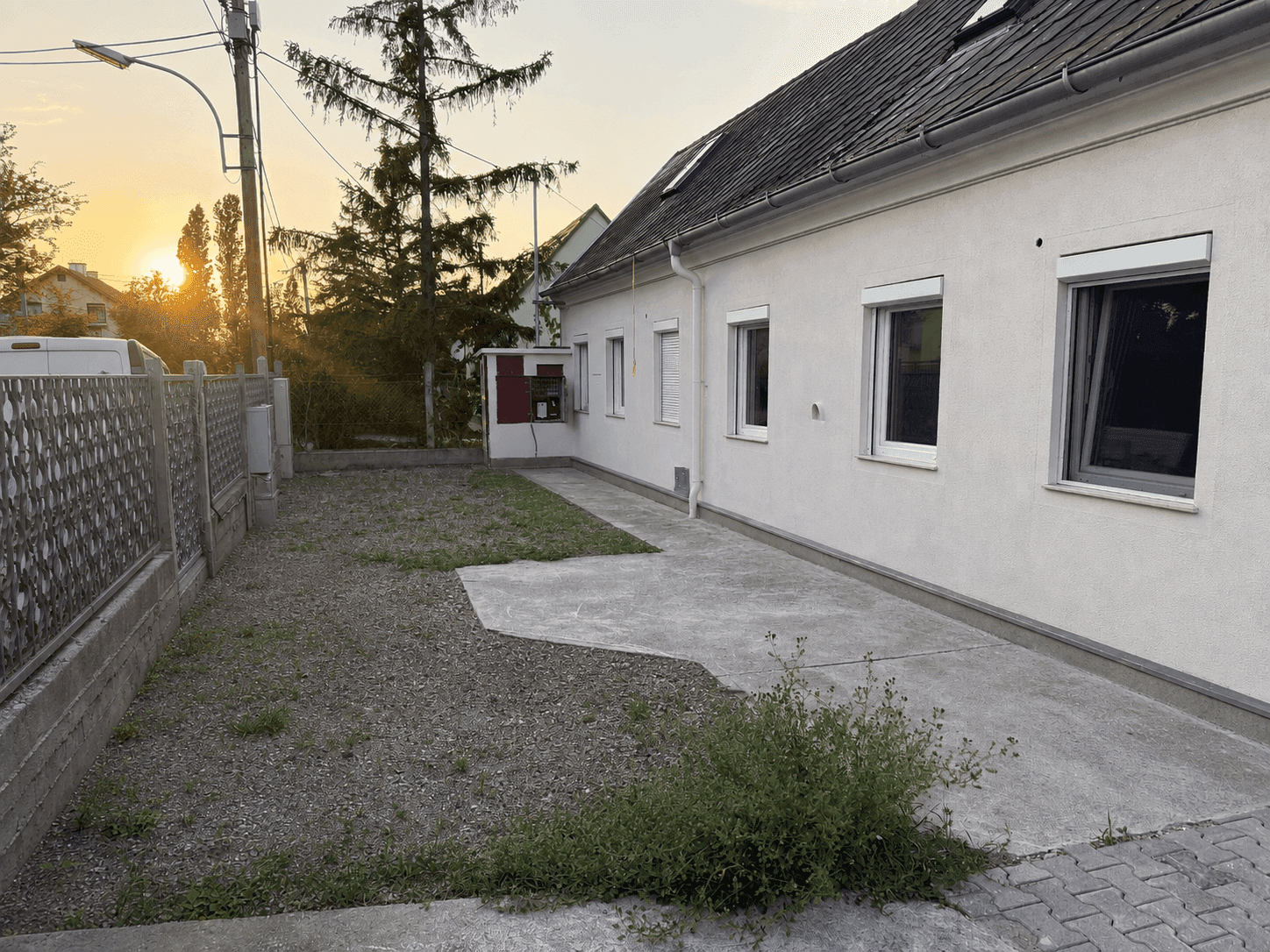 Seitenansicht eines weißen Hauses mit Kies- und Betonfläche im Vorgarten bei Sonnenuntergang.