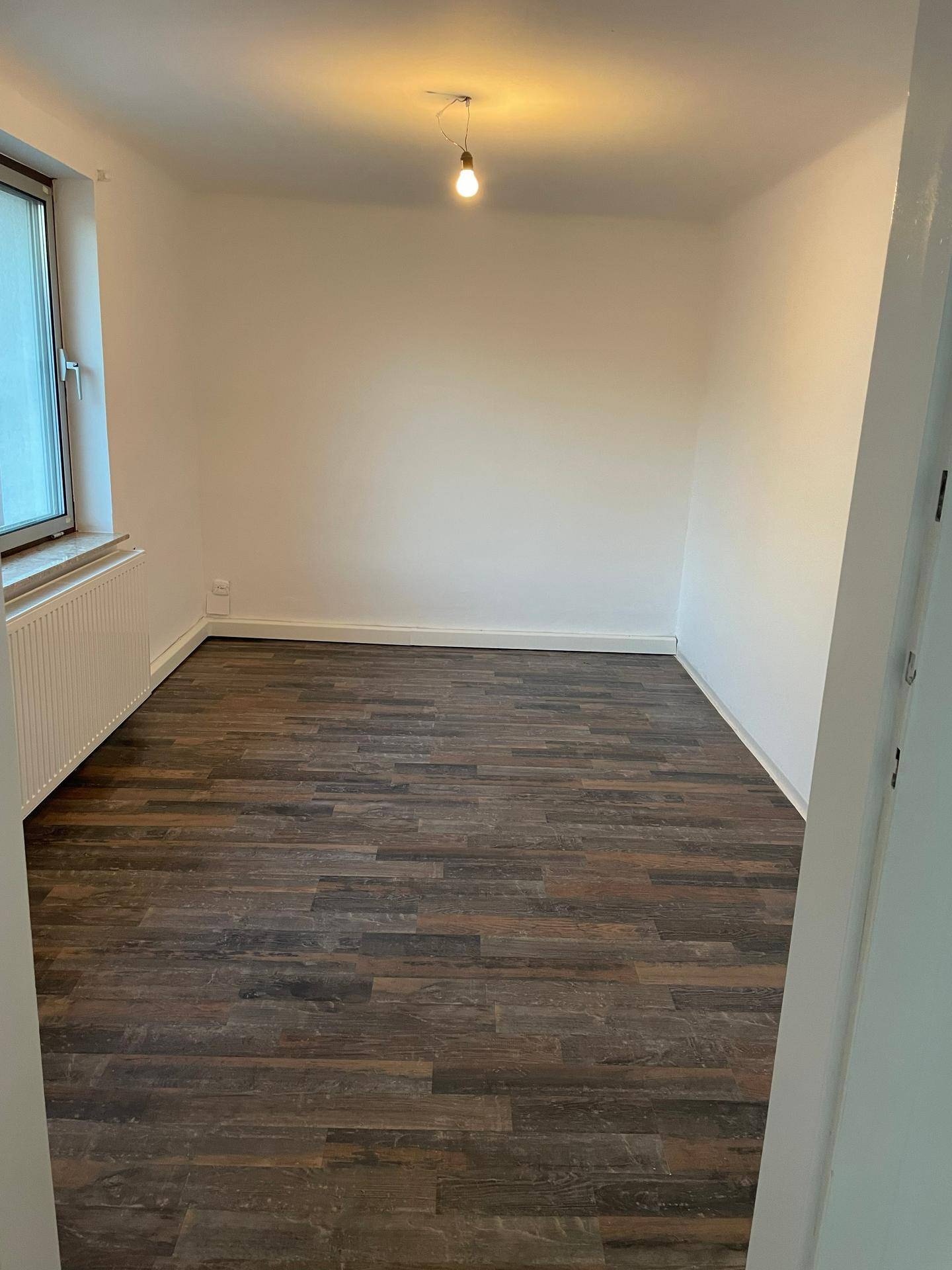 Helles Schlafzimmer mit dunklem Laminatboden, weißen Wänden und einem Fenster mit Heizkörper.