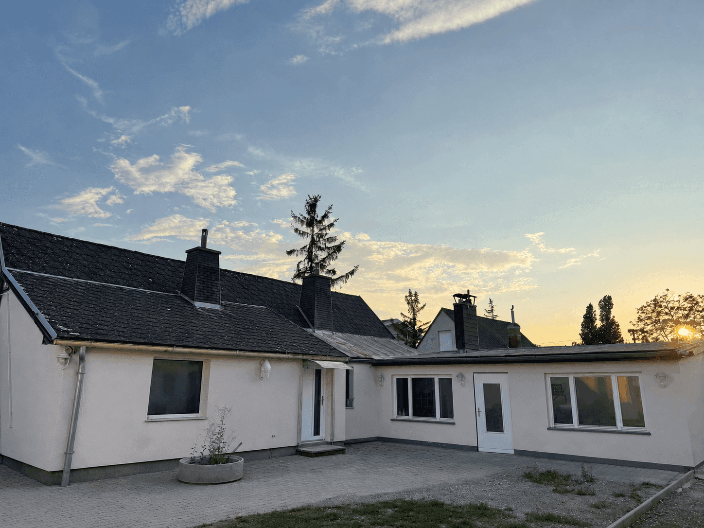 Rückansicht eines weißen Hauses mit dunklem Dach, gepflastertem Bereich und Rasenfläche im Abendlicht.