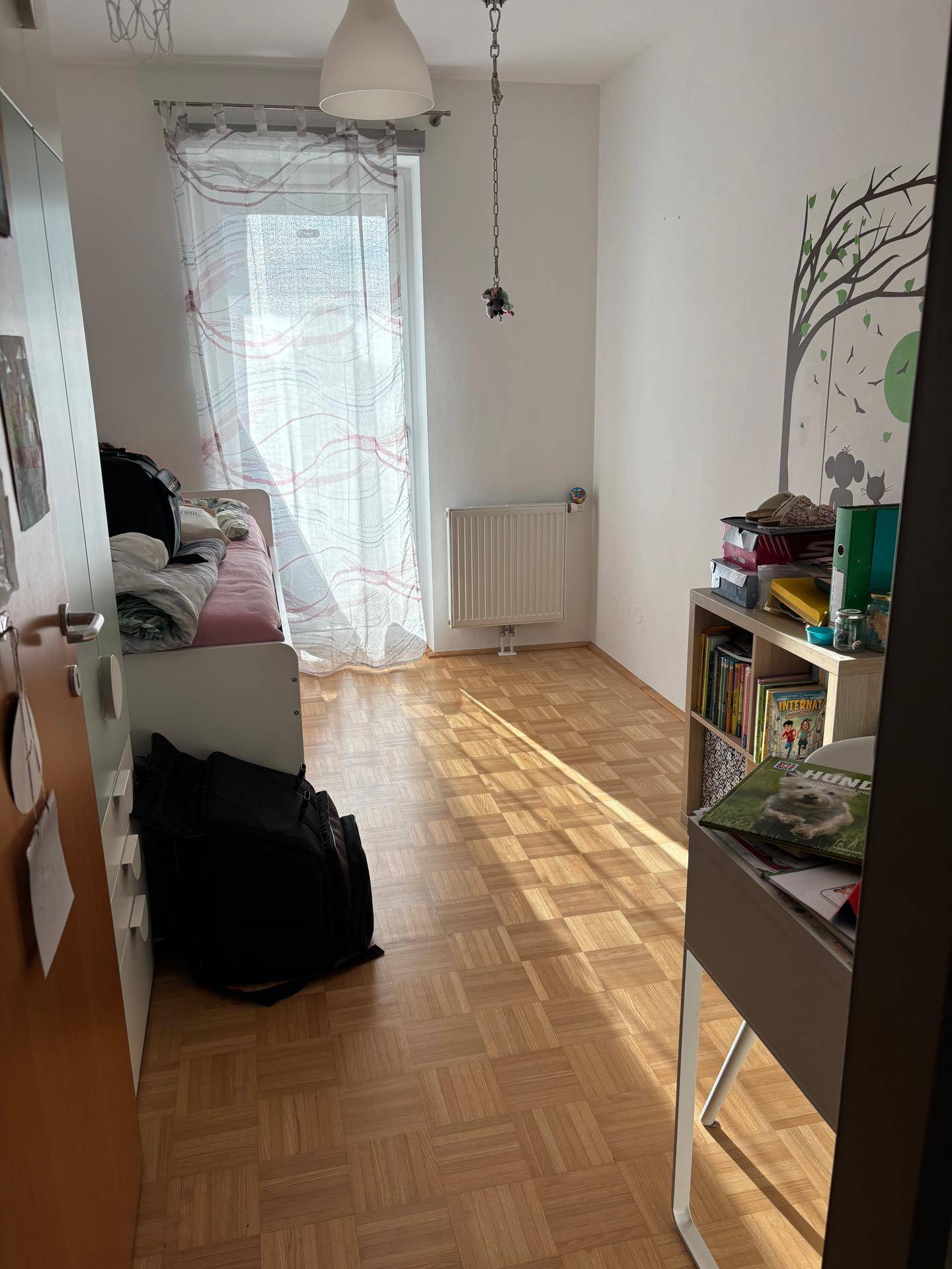 Helles Schlafzimmer mit Parkettboden und einem großen Fenster, das viel Tageslicht hereinlässt.