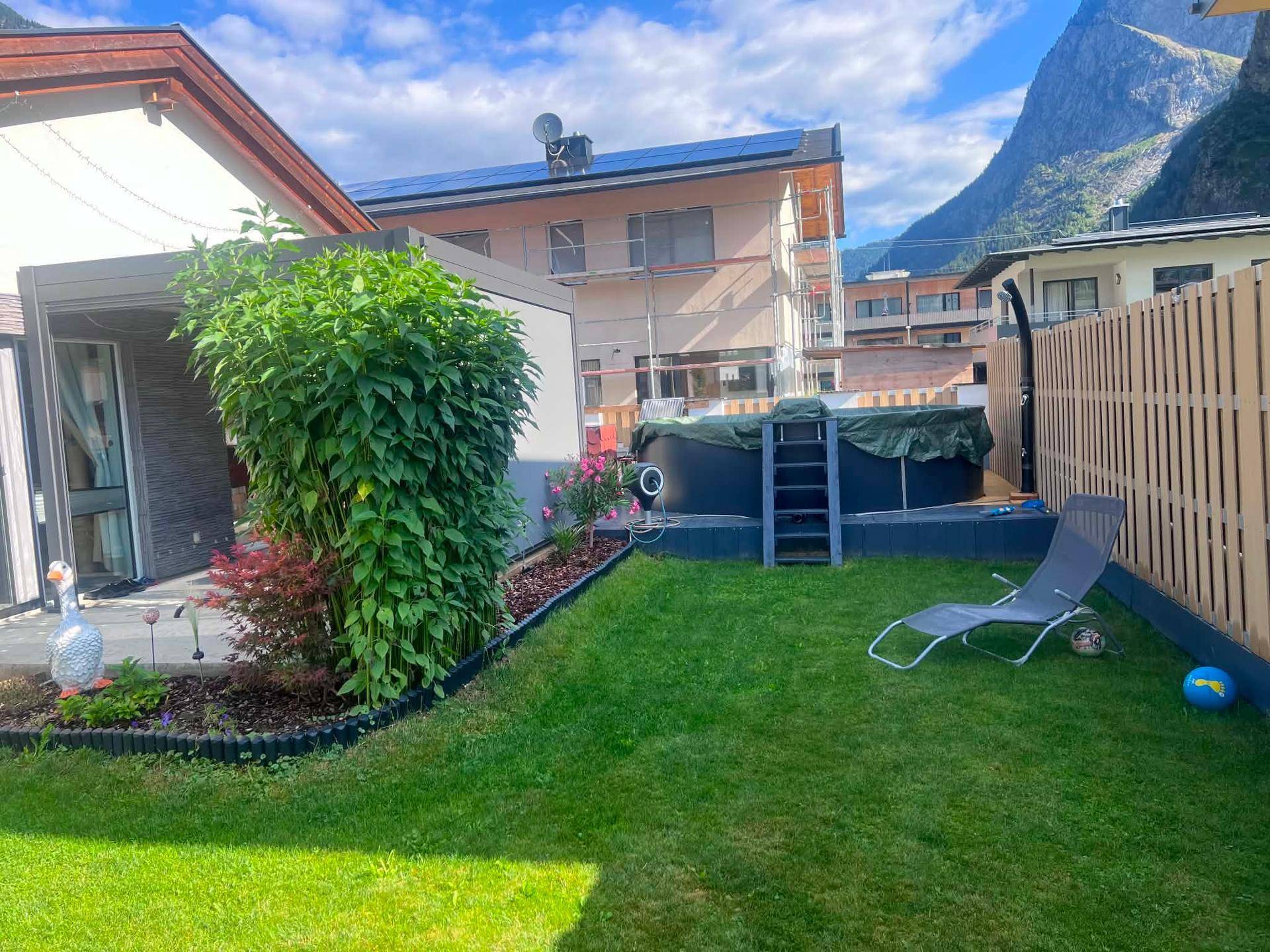Gepflegter Garten mit Rasenfläche, einem Aufstellpool und Liegestuhl vor beeindruckender Bergkulisse.