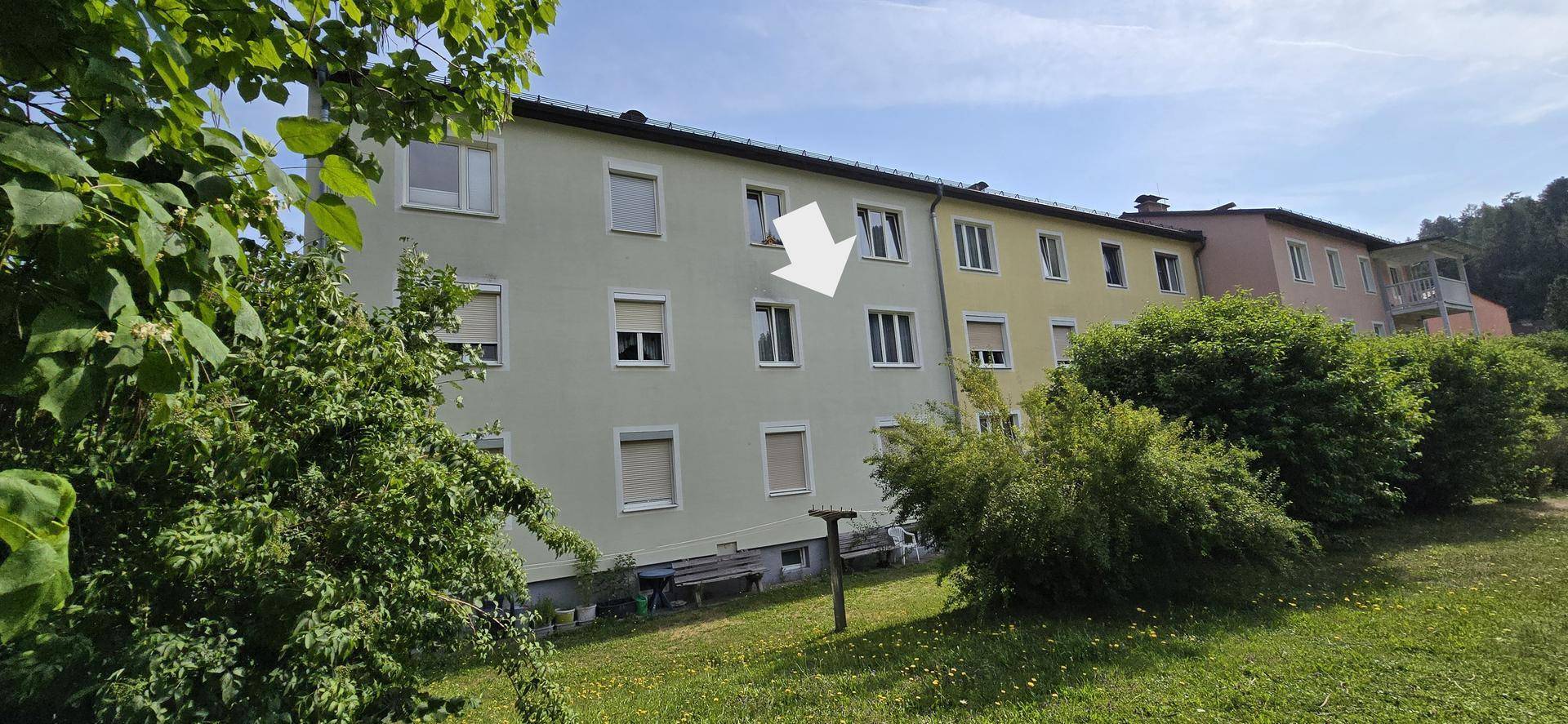 Mehrfamilienhaus mit heller Fassade, Balkonen und grüner Umgebung unter blauem Himmel.