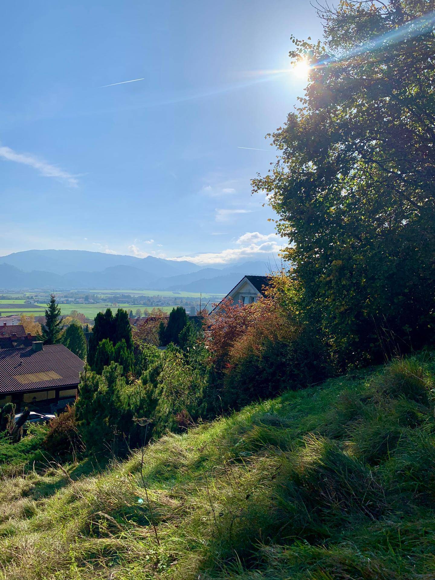 Sonniger Ausblick über ein grünes Tal mit Häusern und Bergen, eingefangen von einem grasbewachsenen Hang.