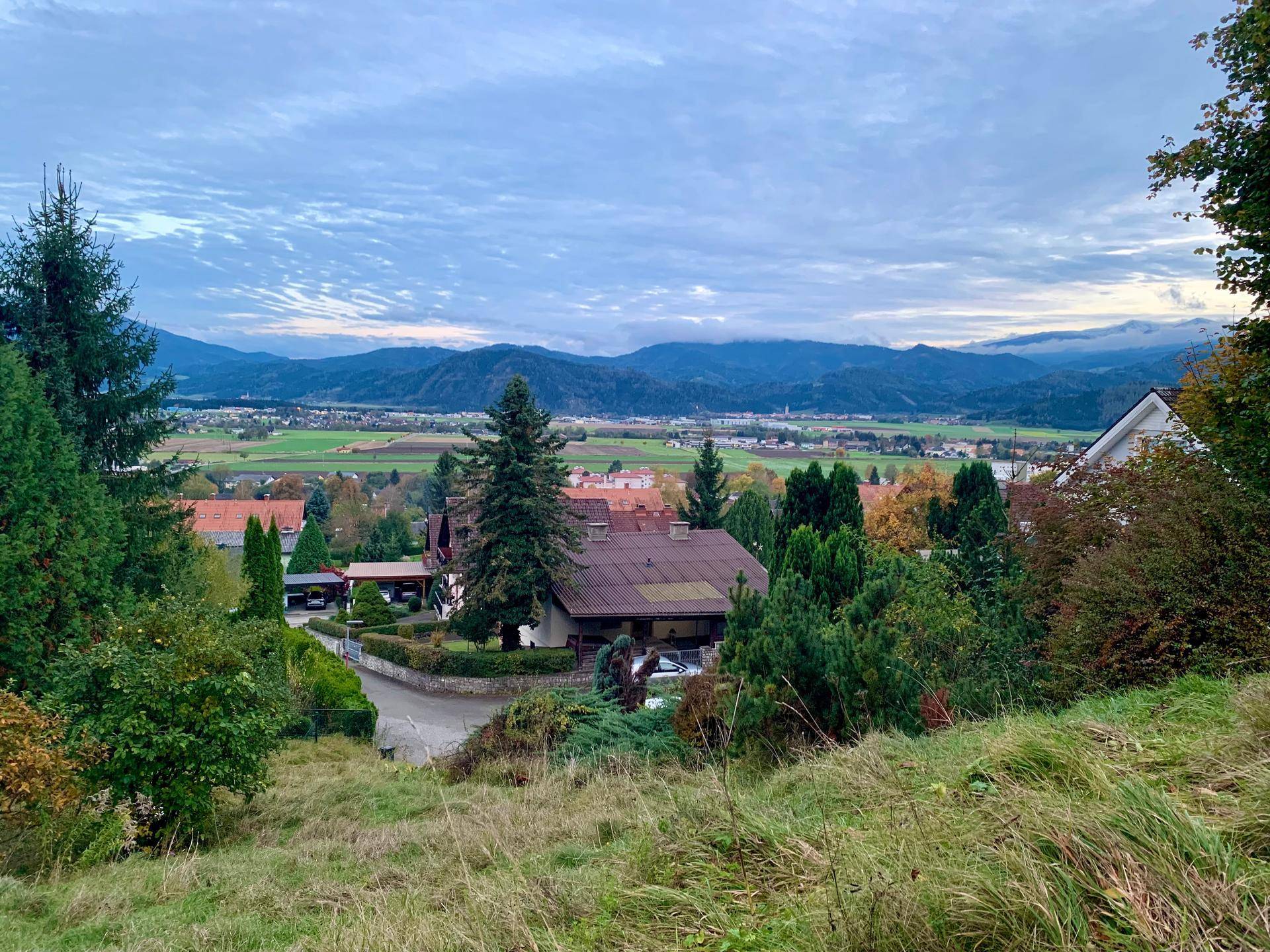 Panoramablick über ein Wohngebiet in einem Tal mit Bergen im Hintergrund unter einem bewölkten Himmel.