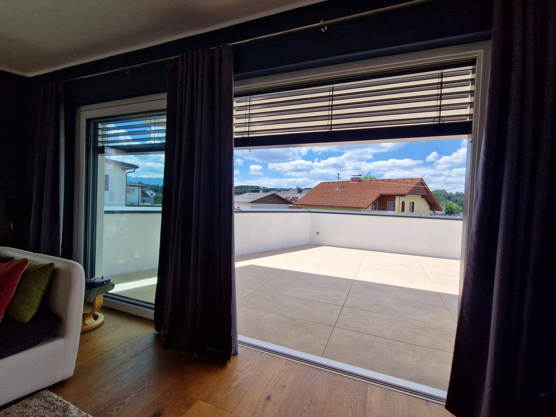 Blick vom Wohnzimmer auf die weitläufige Terrasse und die umliegende Wohngegend mit Bergen im Hintergrund.