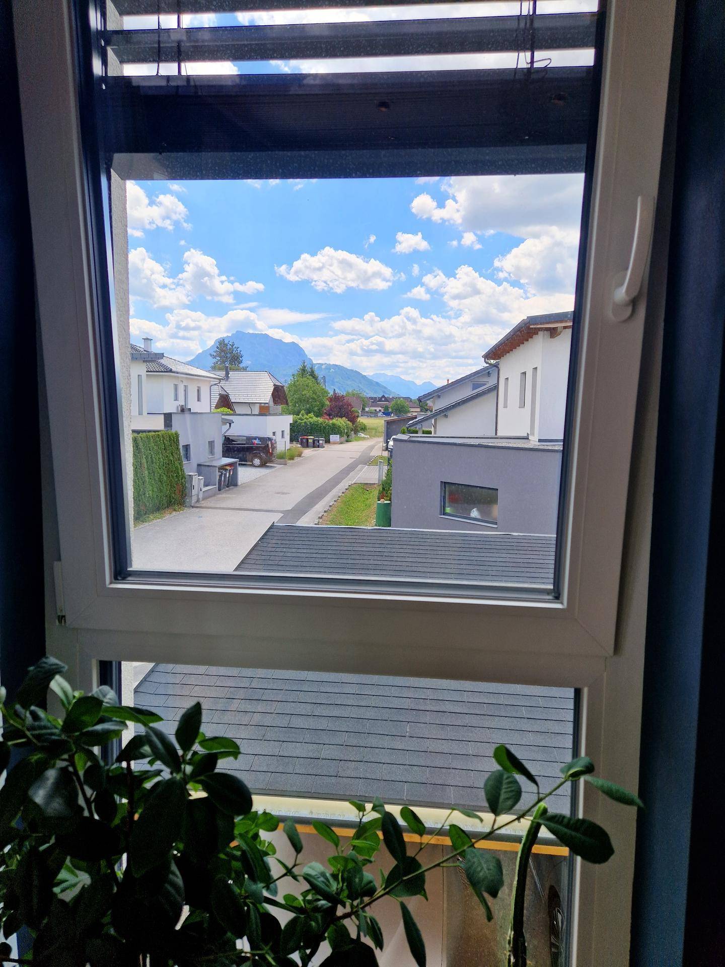 Blick aus dem Fenster auf eine Wohnstraße mit Häusern und Bergen im Hintergrund.