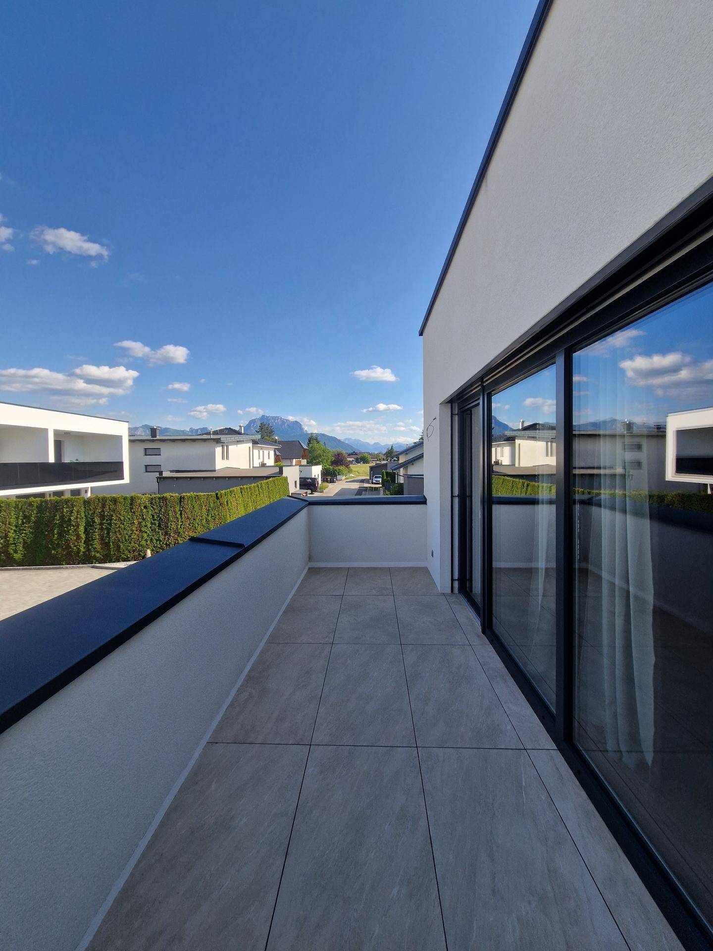 Schmaler Balkon mit Fliesenboden und Blick auf moderne Gebäude sowie die umliegende Berglandschaft.