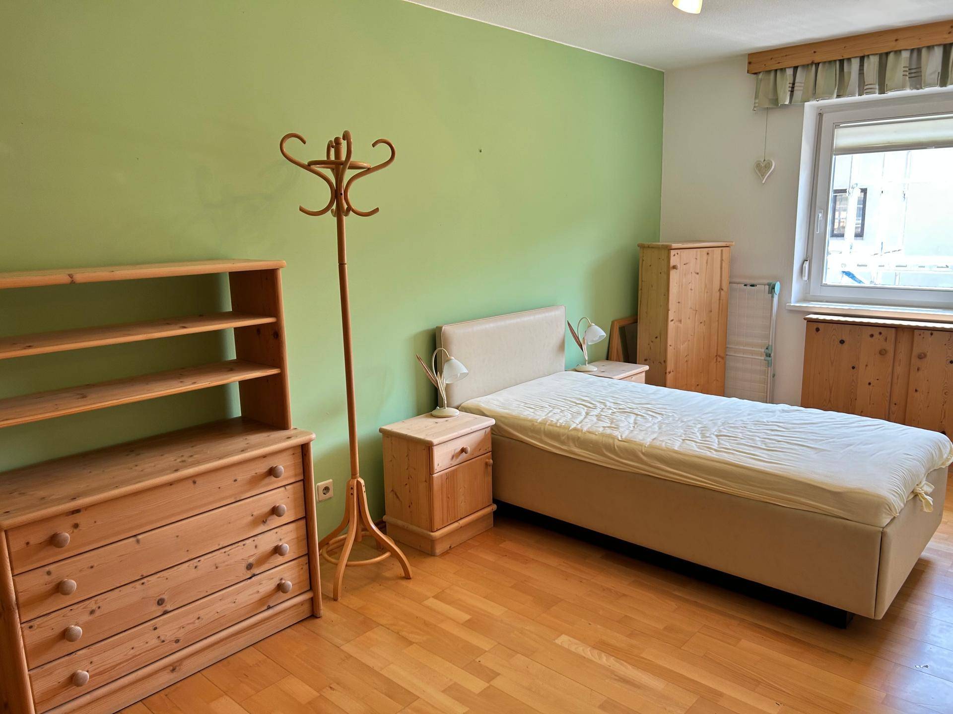 Schlafzimmer mit Einzelbett, Holzkommode und einer auffälligen grünen Akzentwand.
