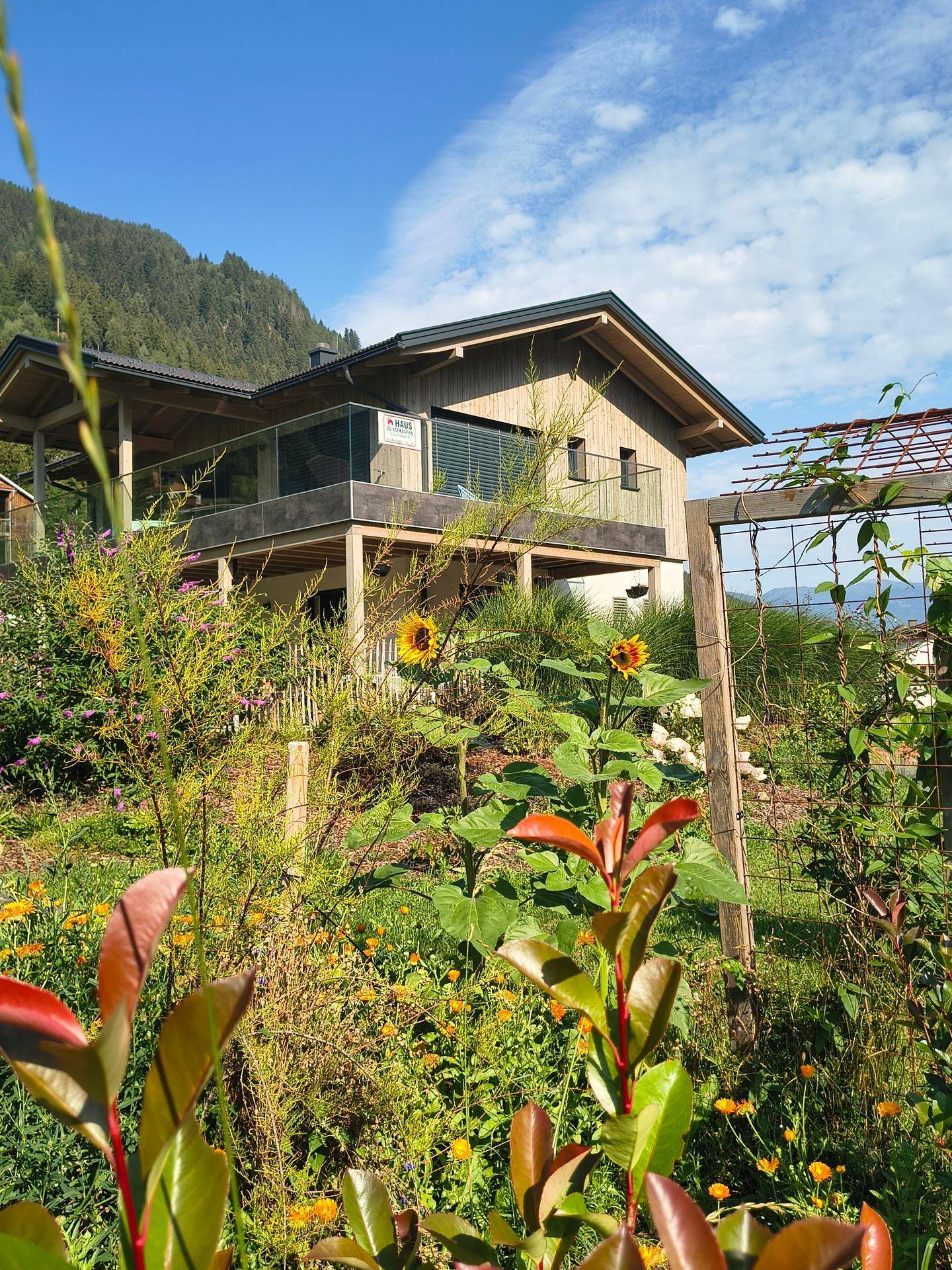 Ansicht des Holzhauses durch blühende Gartenpflanzen, mit Balkon und Blick auf die umliegende Berg- und Seenlandschaft.