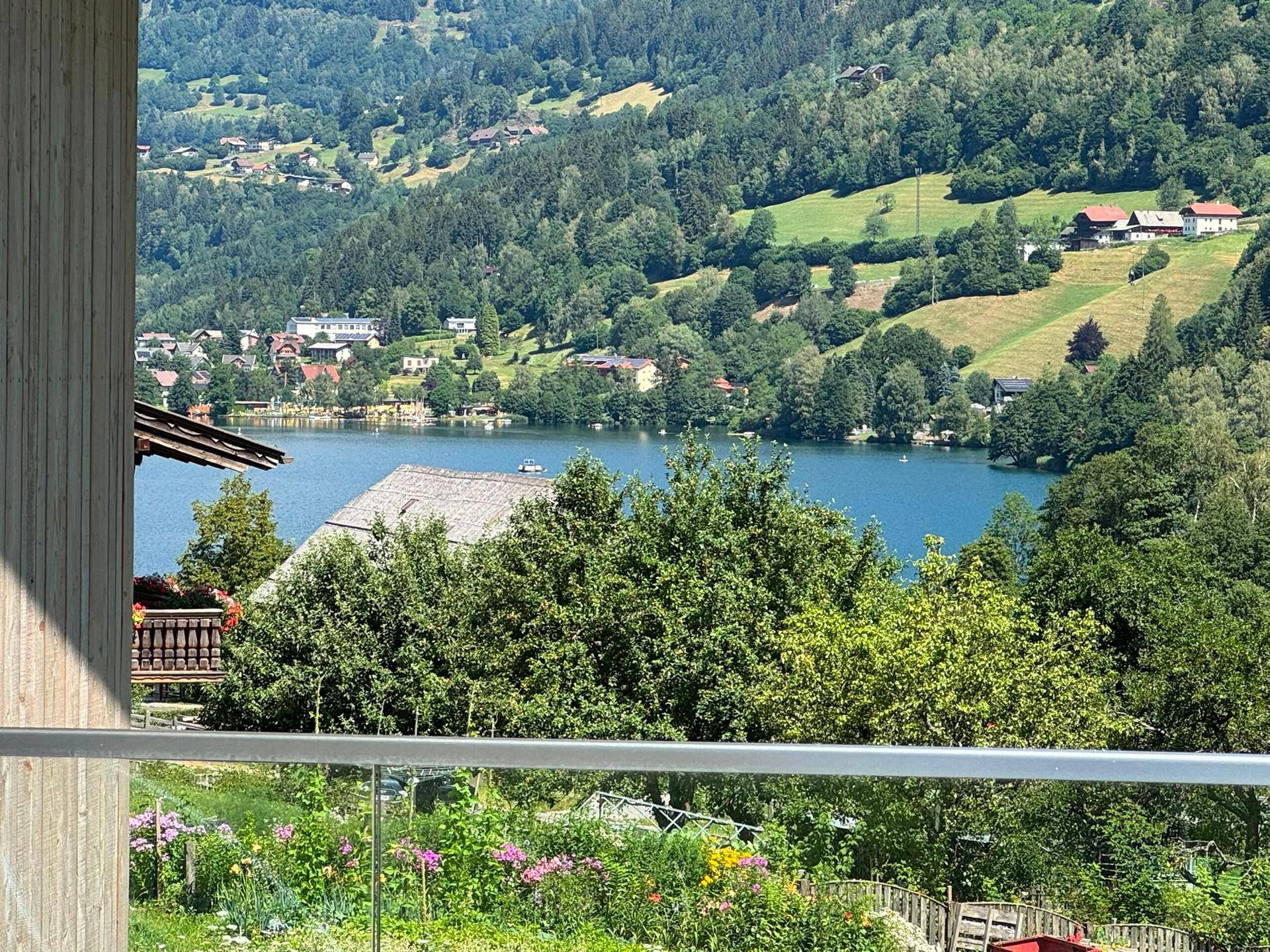 Panoramablick vom Balkon auf den glitzernden See und die majestätischen Berge, umgeben von grüner Natur.