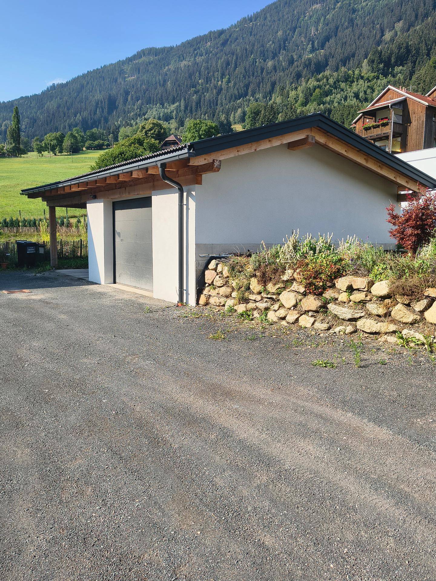 Separate Garage mit Carport und Schotterauffahrt, umgeben von einer Steinmauer und grüner Landschaft.