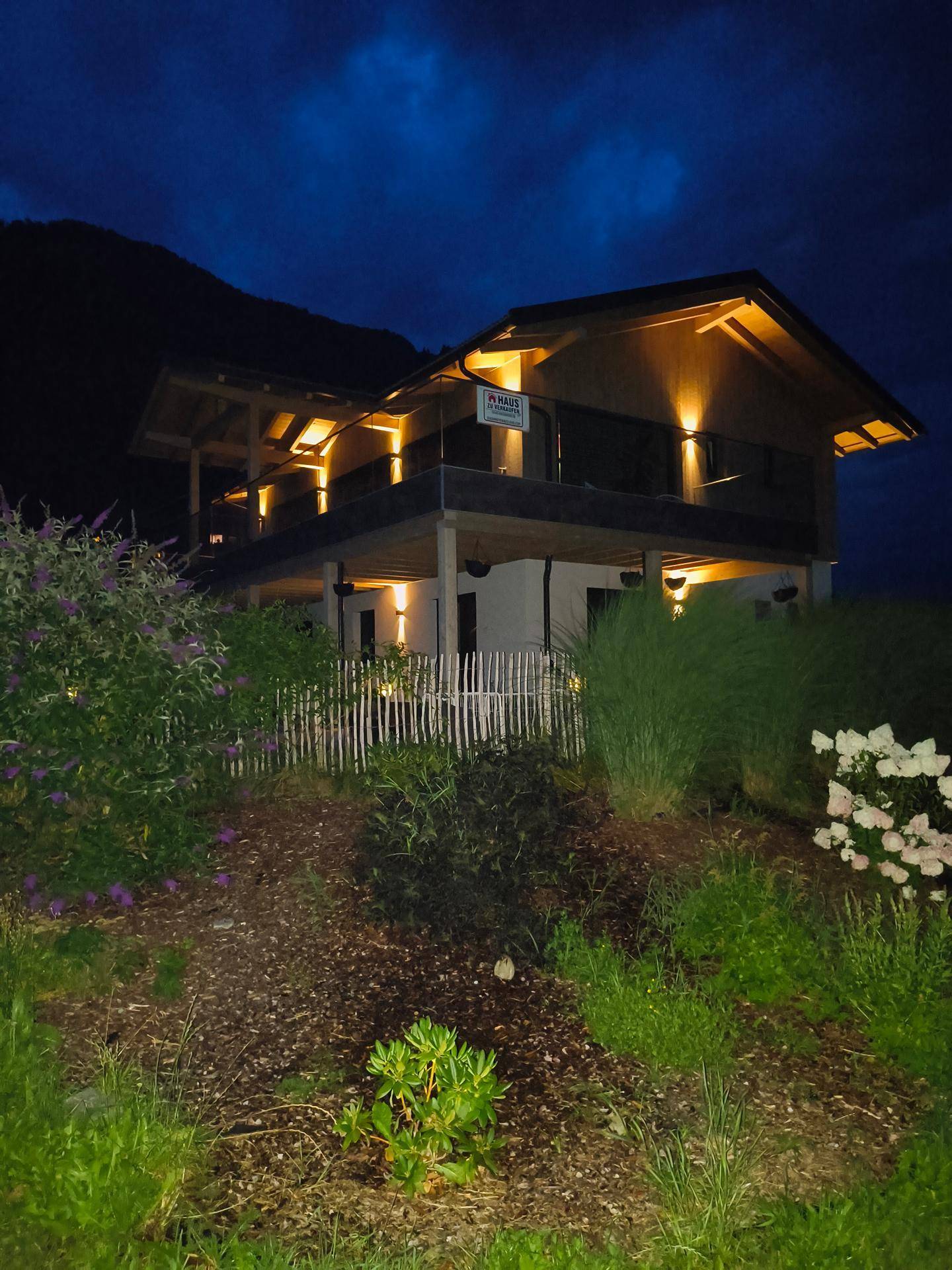 Das moderne Haus bei Nacht, beleuchtet mit warmem Licht, umgeben von üppiger Vegetation und dunklem Nachthimmel.