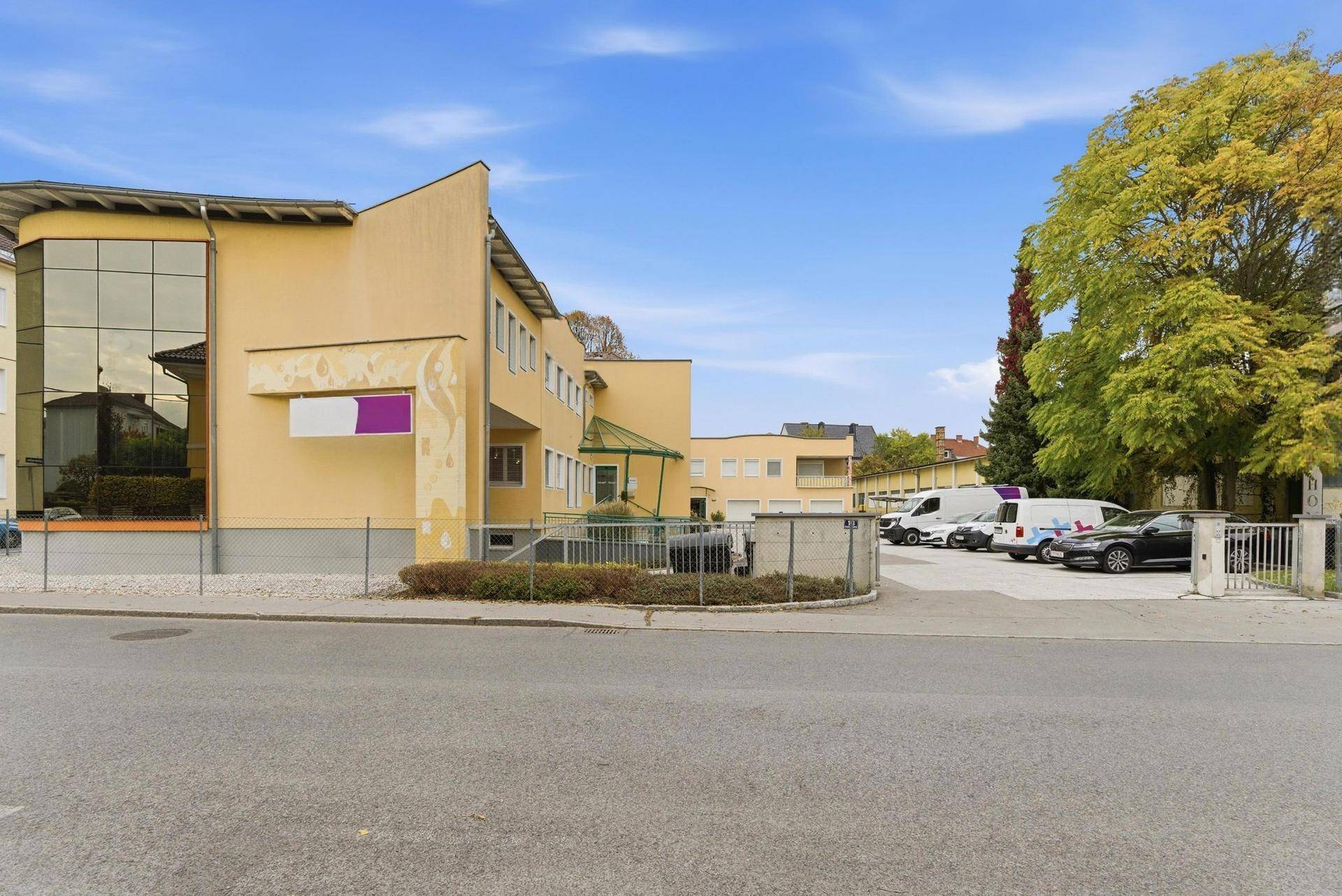 Seitenansicht des gelben Gebäudes mit Glasfassade, Eingangsbereich und Außenparkplätzen.