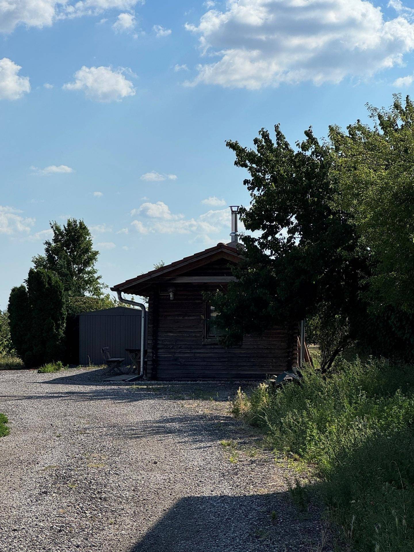 Holzhütte mit angrenzendem Schuppen und Kiesweg, umgeben von Bäumen und Sträuchern.