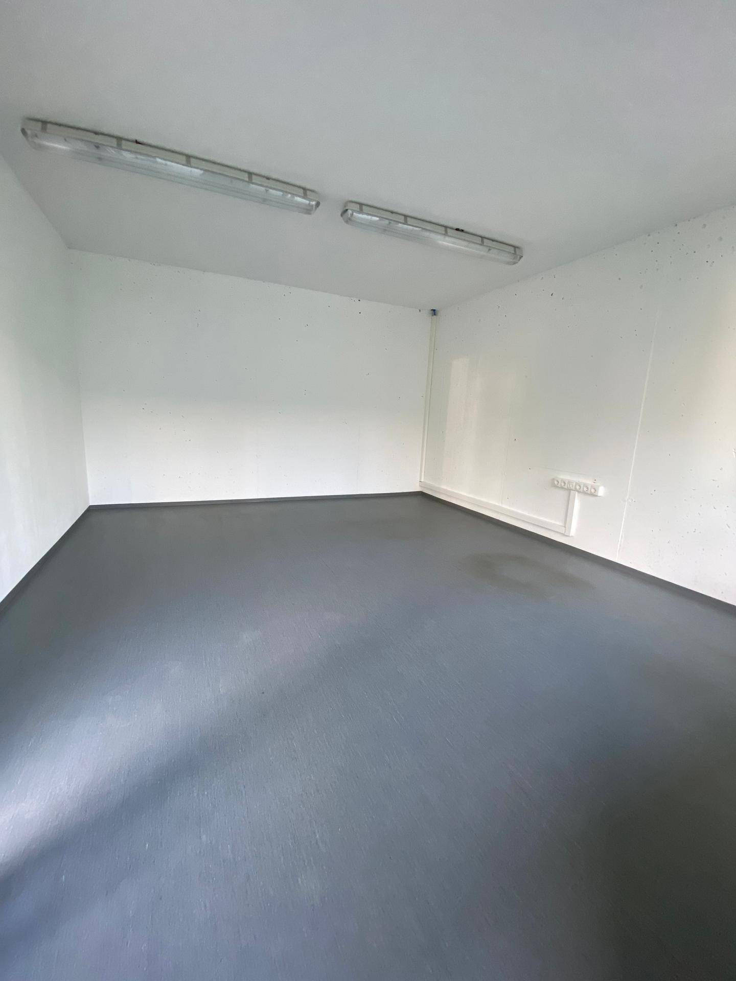 Kleiner, leerer Raum mit weißen Wänden und einem dunklen Boden, beleuchtet von Deckenleuchten.