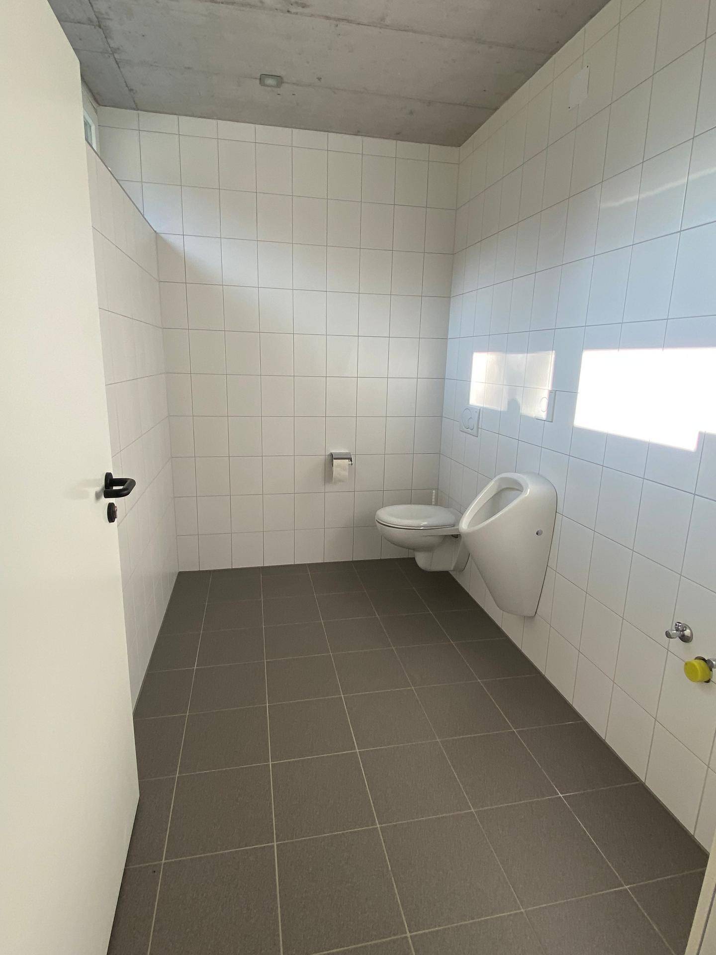Modernes Badezimmer mit weißen Fliesen, Toilette, Urinal und grauen Bodenfliesen, gut beleuchtet.