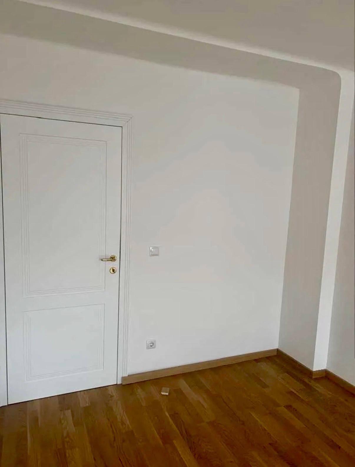 Ecke eines leeren Zimmers mit Parkettboden und weißer Tür, die zu einem weiteren Raum führt.