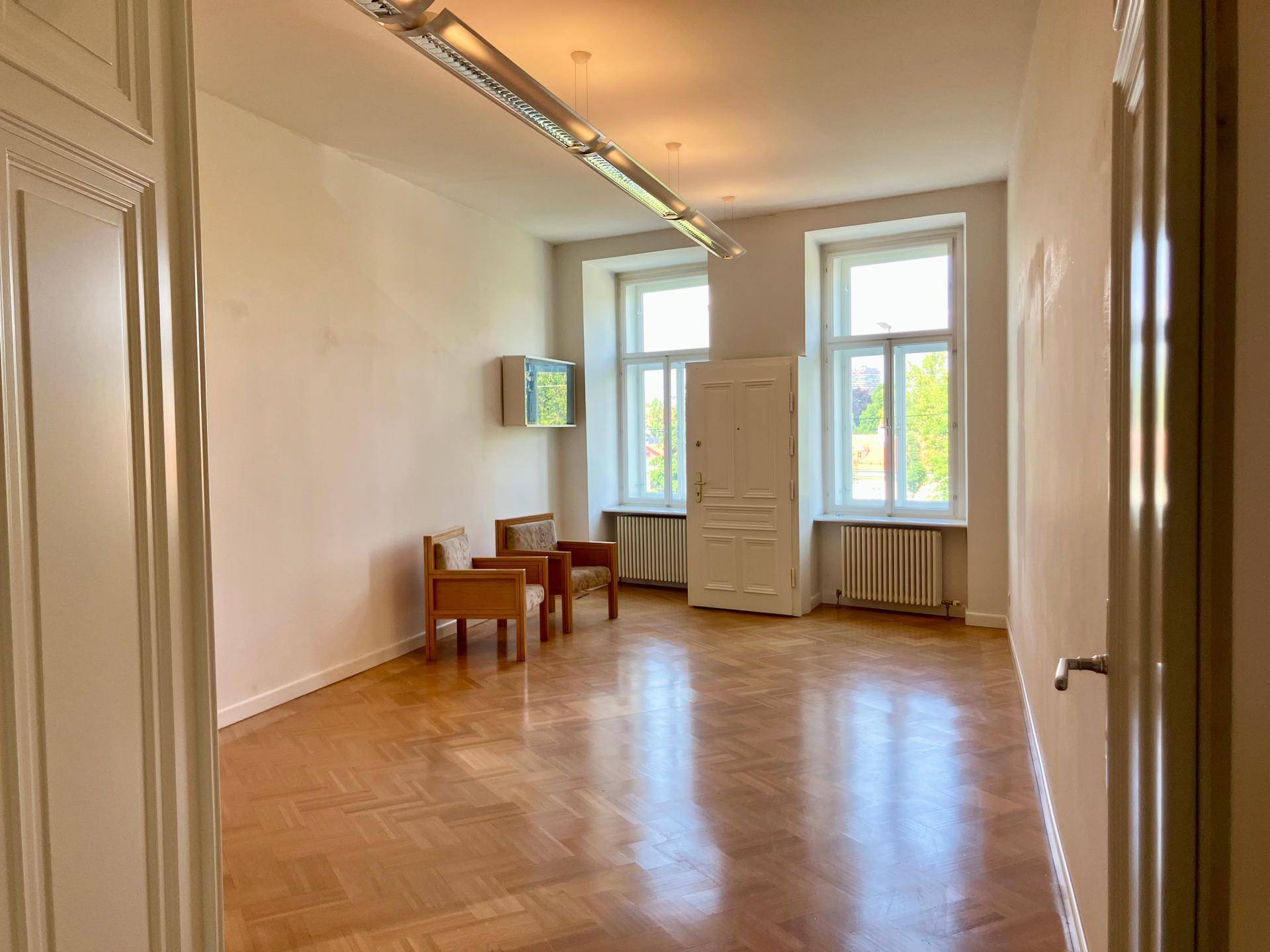 Leerer Büroraum mit Parkettboden, zwei Fenstern und zwei Sesseln, ideal als Wartebereich oder kleines Büro.