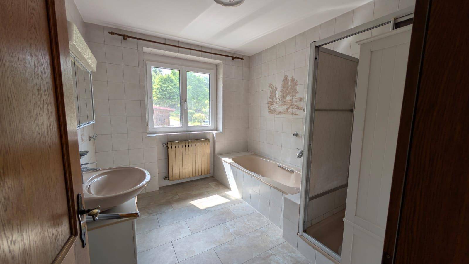 Helles Badezimmer mit Badewanne, Dusche und Fenster, gefliest in hellem Beige.