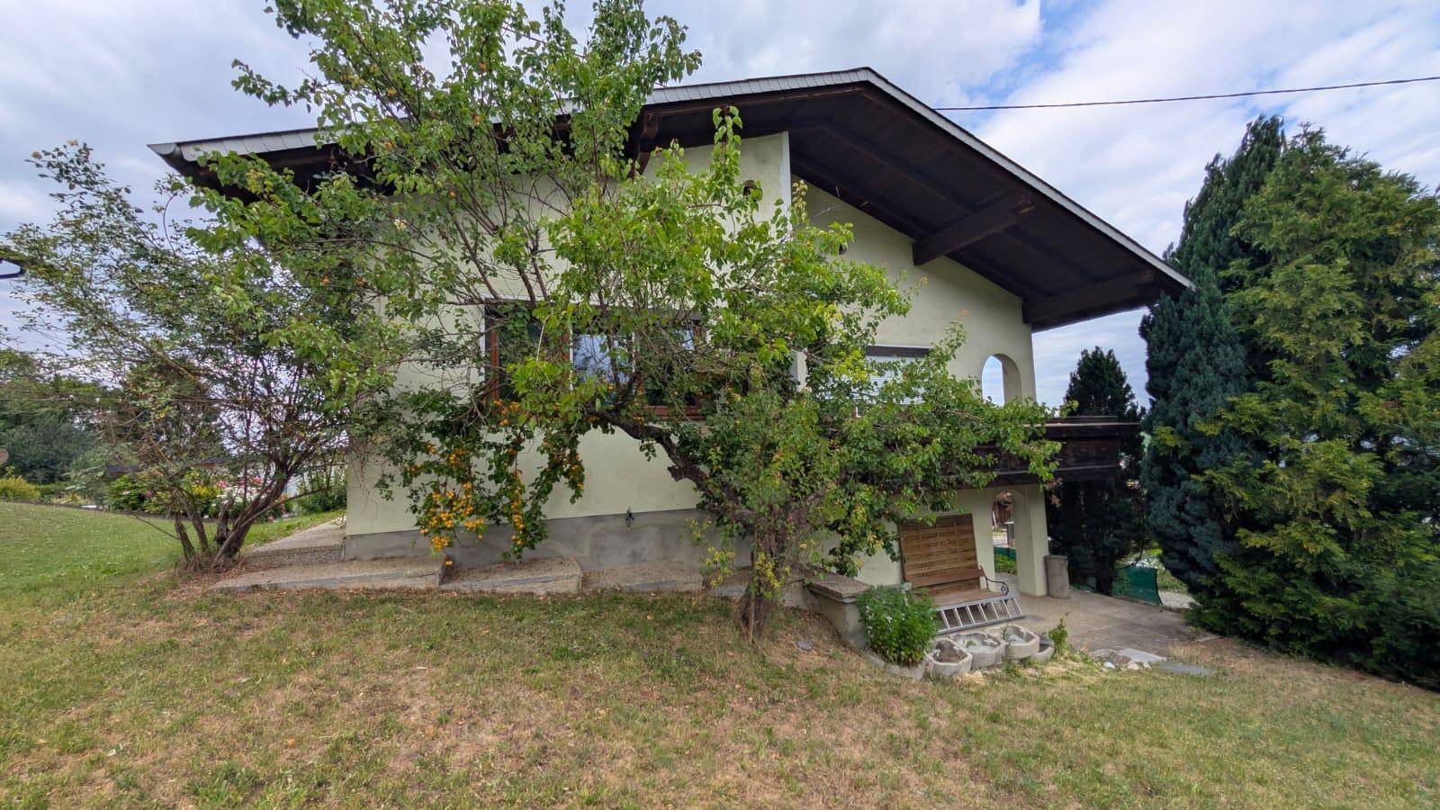 Hausansicht von der Seite mit Balkon und üppiger Vegetation im Gartenbereich.