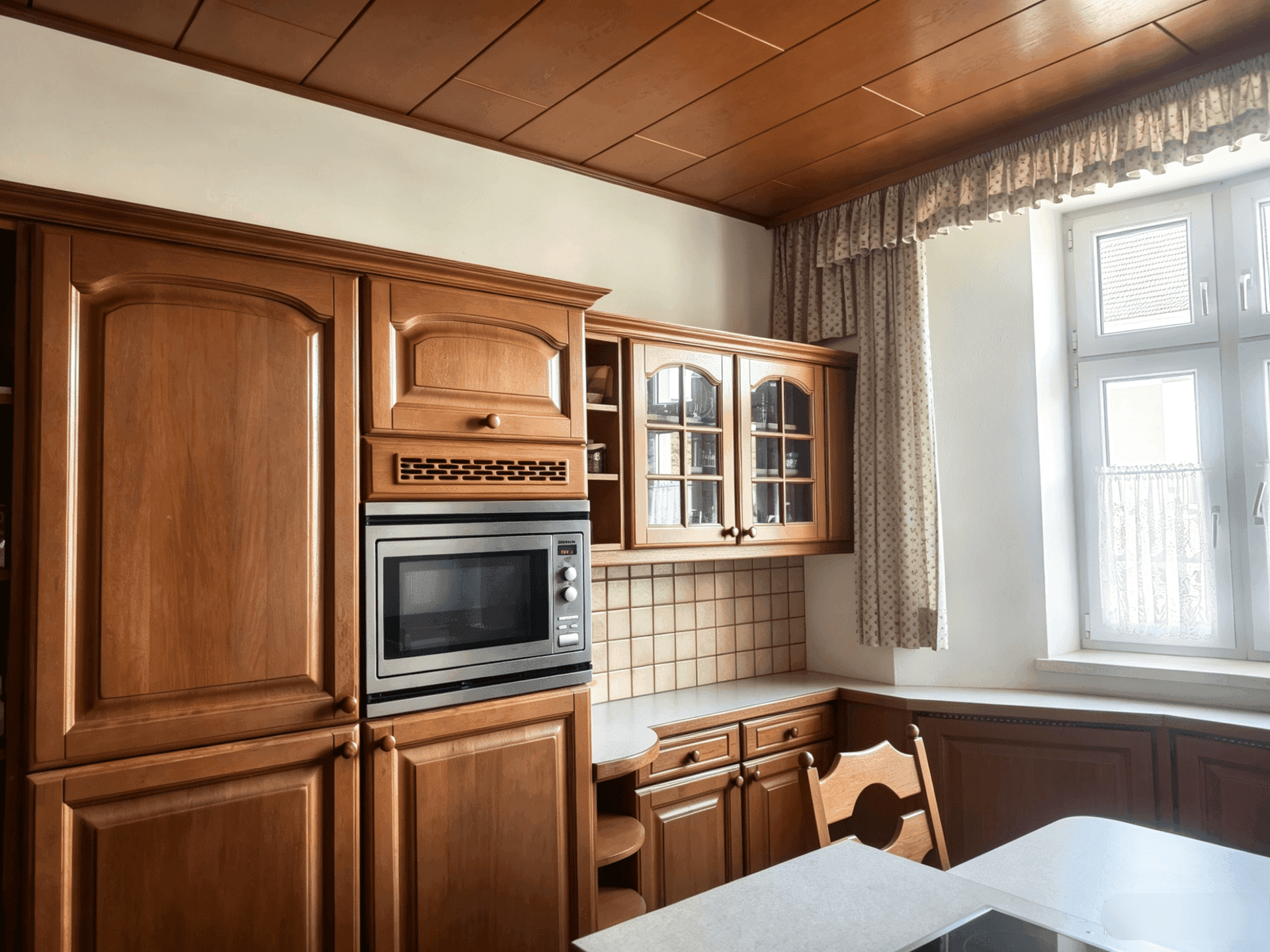Traditionelle Küche mit Holzschränken, eingebauter Mikrowelle und einem Fenster mit Gardinen.