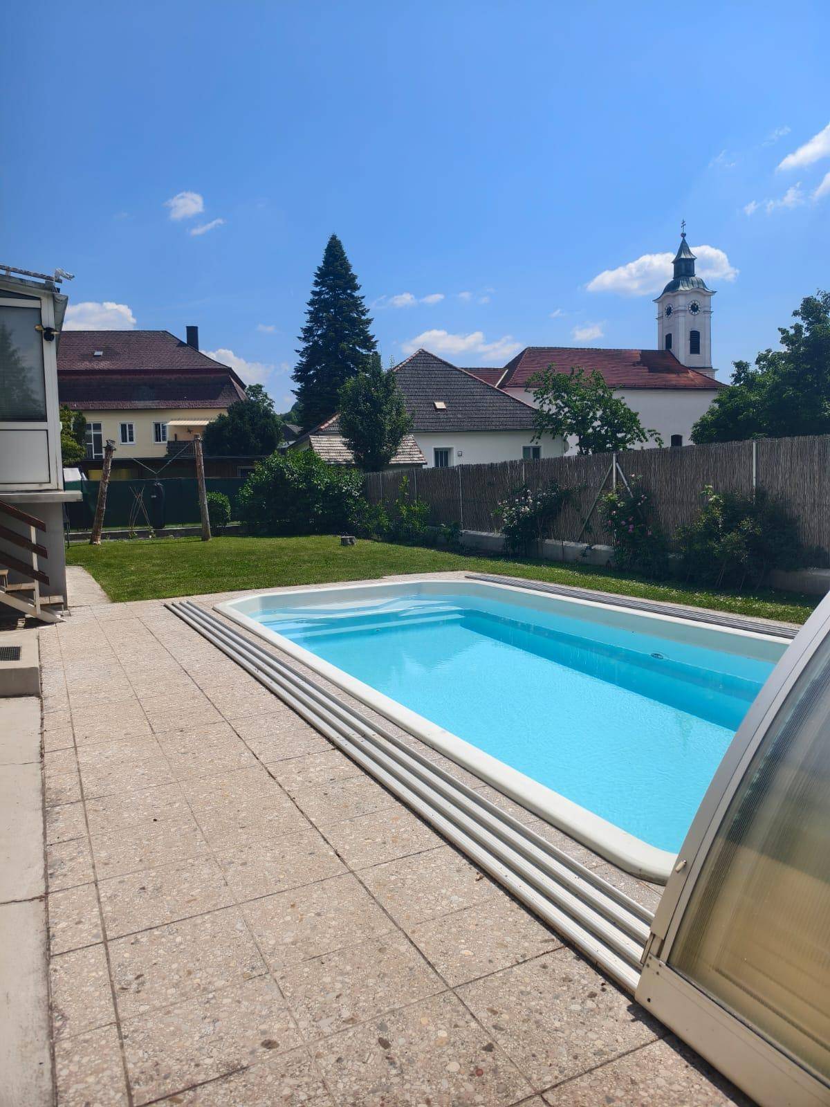 Außenpool mit Überdachung, umgeben von einer gepflasterten Terrasse und Rasenfläche mit Kirchenblick.