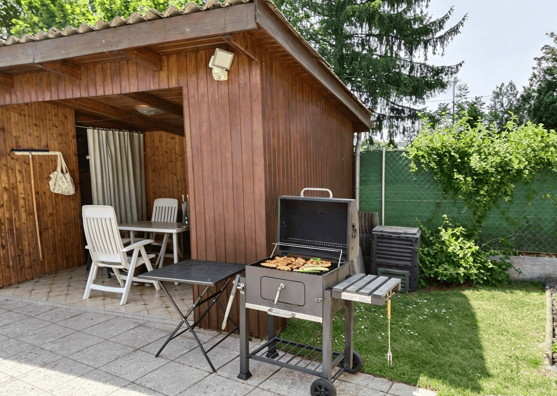 Gemütlicher Grillbereich im Garten neben einem Holzschuppen mit Sitzgelegenheiten im Freien.