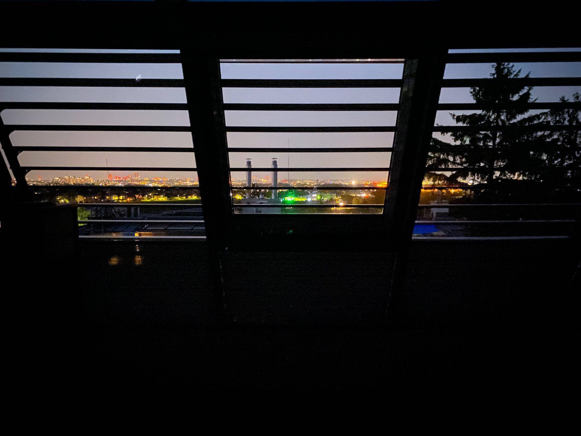 Nächtlicher Ausblick durch ein Fenster mit Jalousien auf die beleuchtete Stadt.