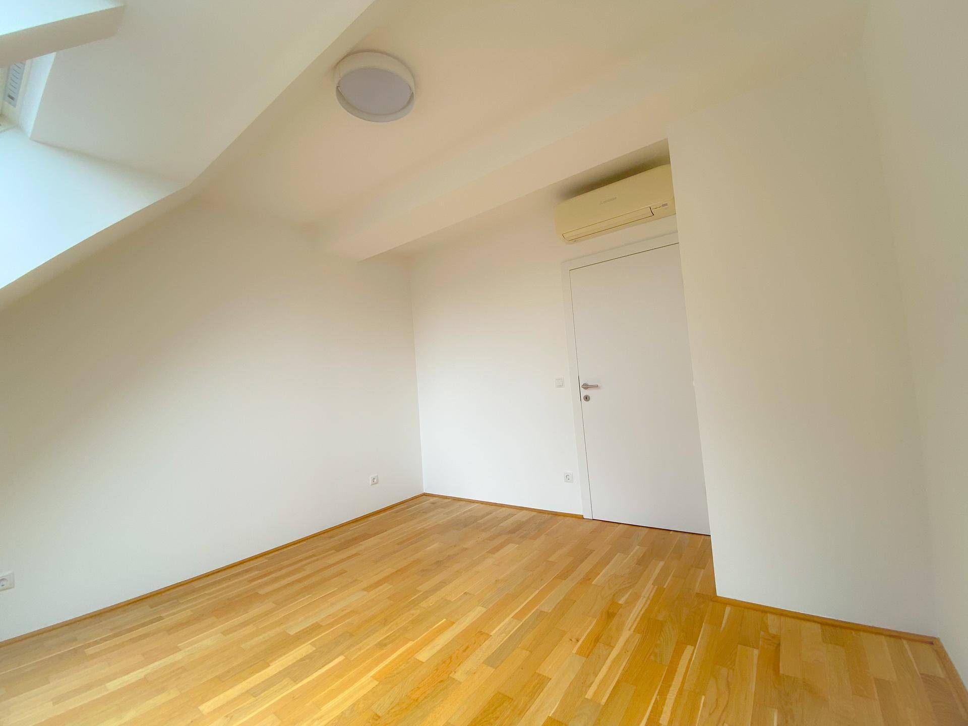 Helles Zimmer im Dachgeschoss mit Holzboden, schräger Decke und Klimaanlage.